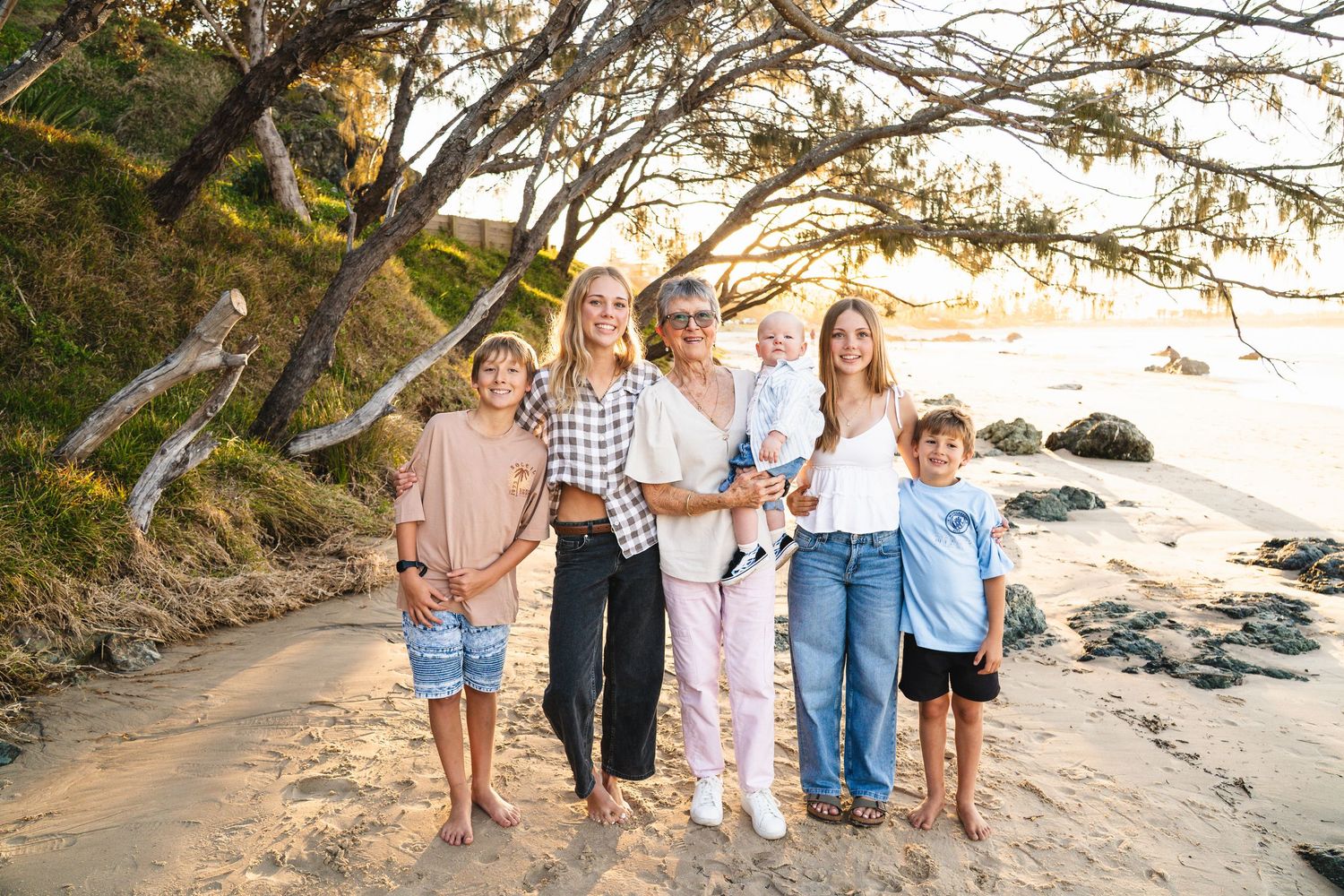 Family of six posing on a sandy beach at golden hour beneath a tree, smiling together near rocky shoreline.