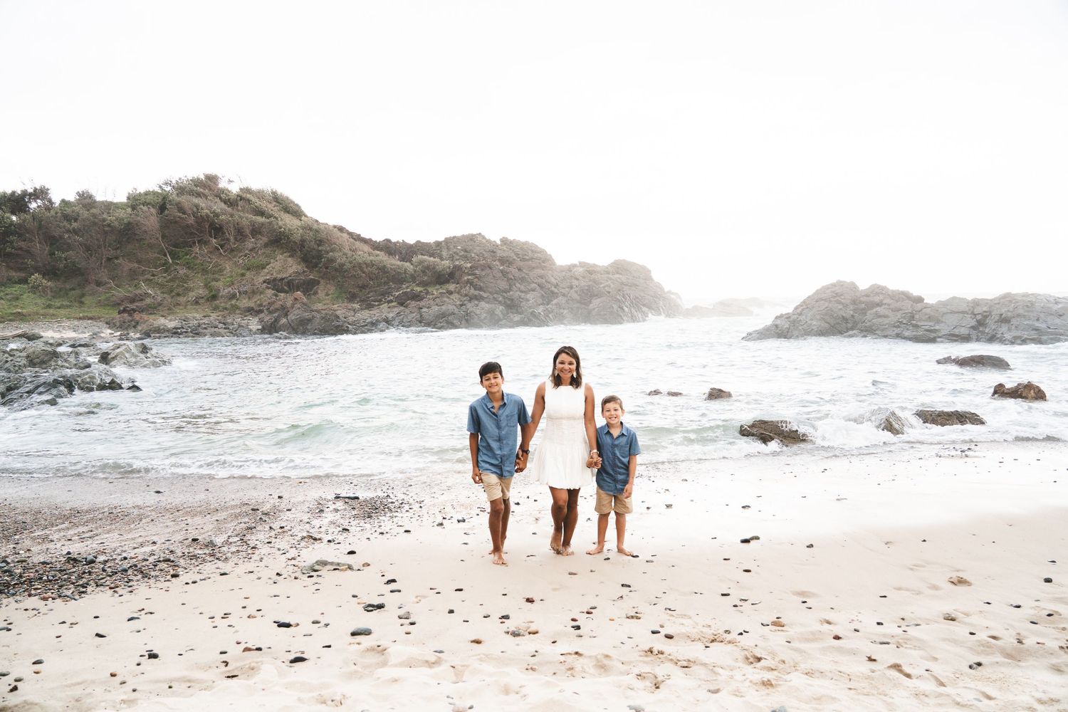 A mother and two children walk hand-in-hand on a sandy beach with rocky cliffs and calm ocean waters behind them.