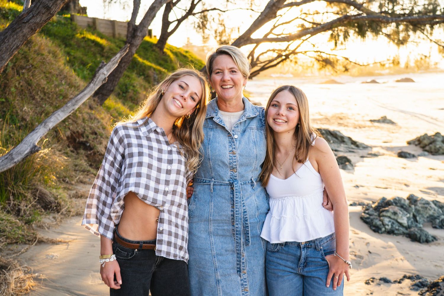 Three women smiling together on a sandy beach at golden hour with rocky shoreline and trees in background.