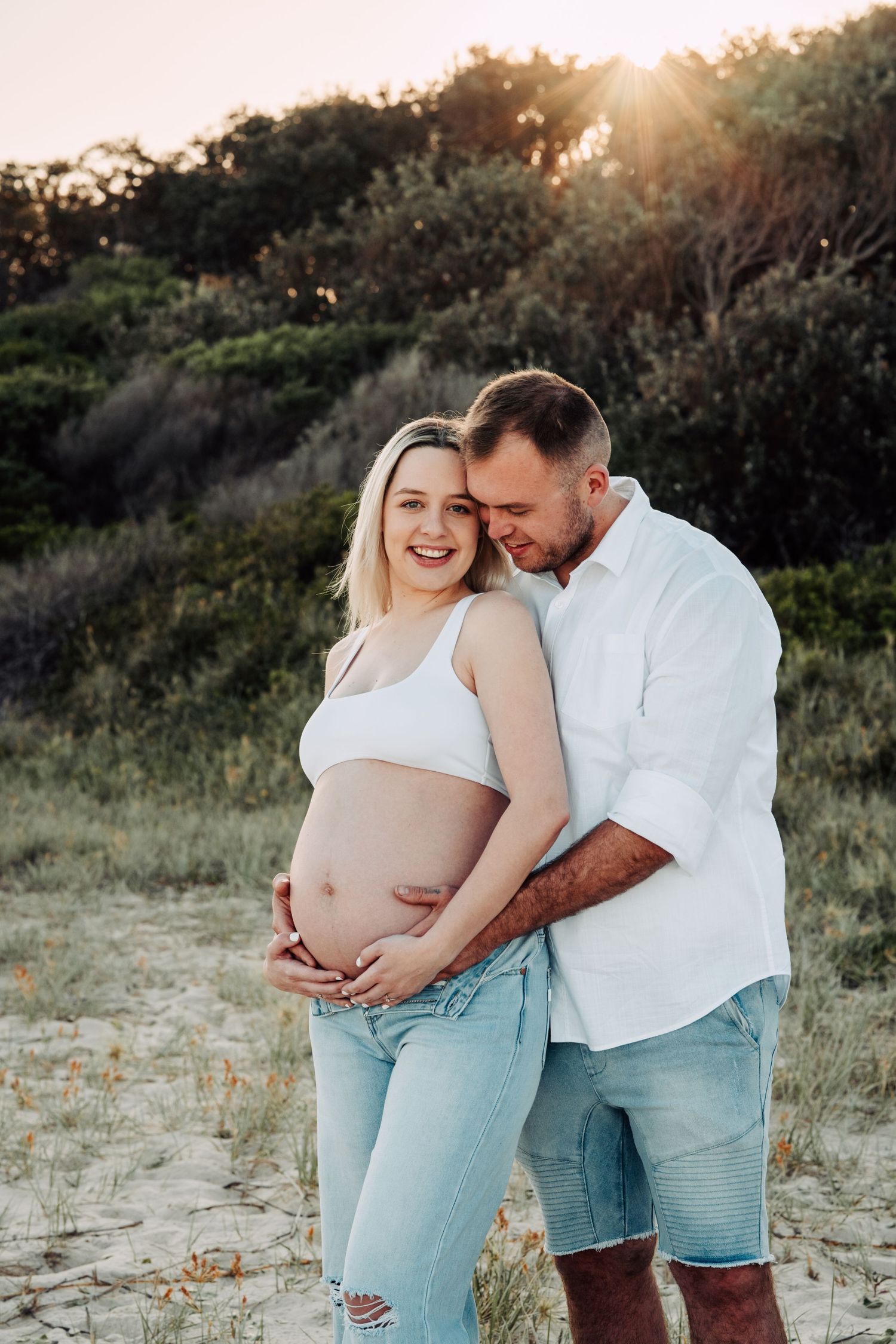 Couple sharing tender maternity moment at sunset on beach, woman smiling with exposed pregnant belly.