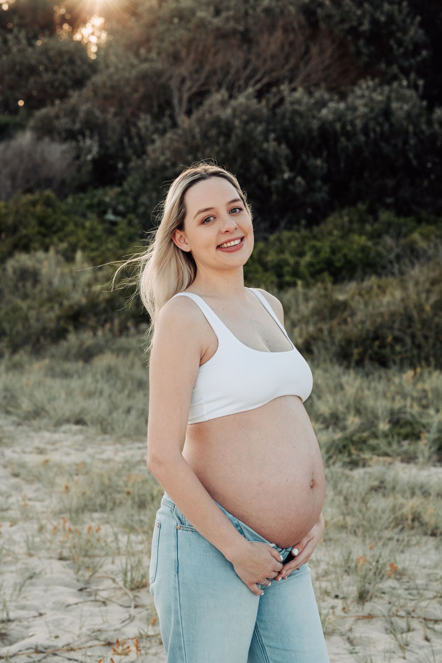Smiling pregnant woman in white crop top and jeans cradling her baby bump on a sandy beach.
