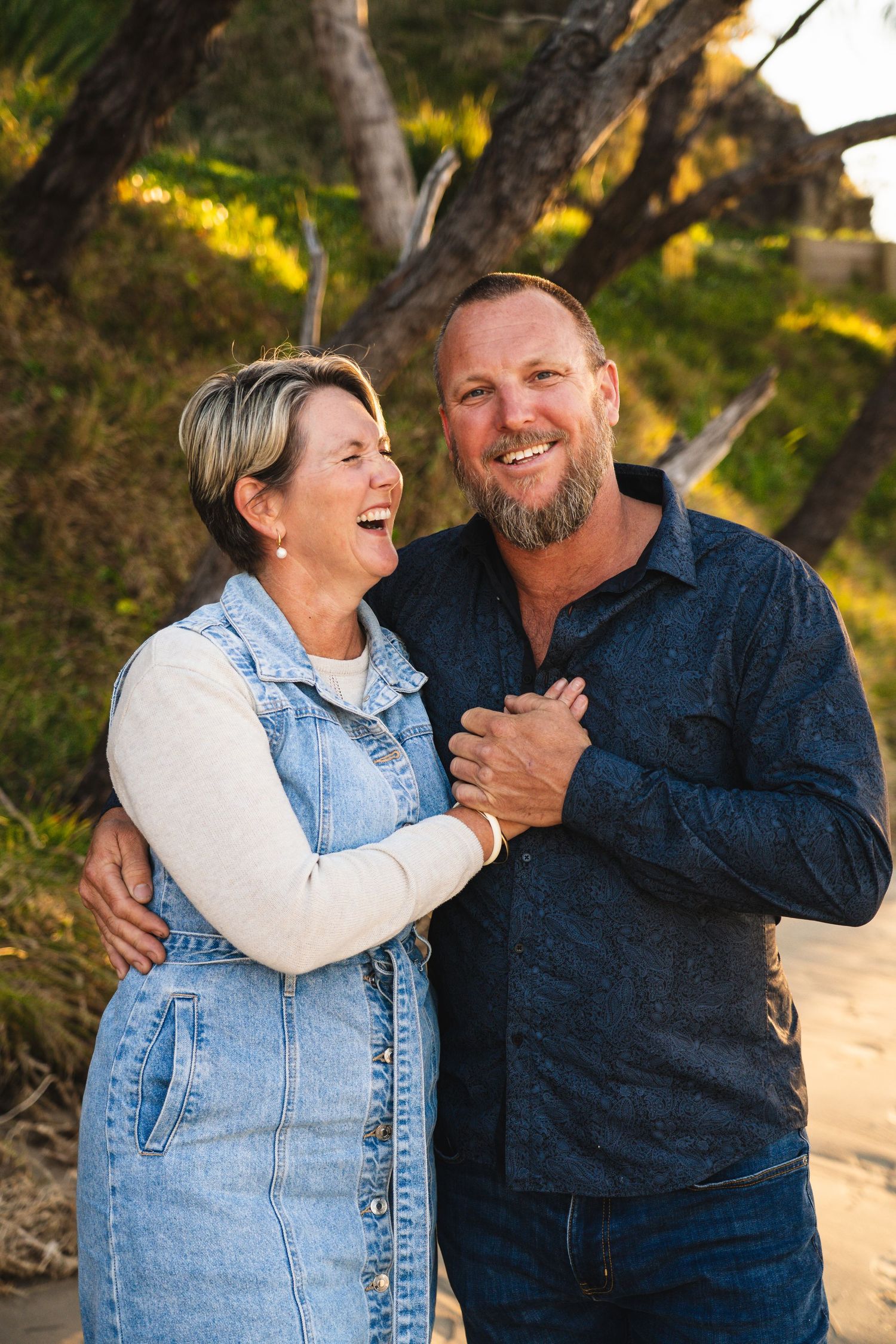 Happy middle-aged couple laughing and embracing outdoors on sandy path with lush green trees at sunset.