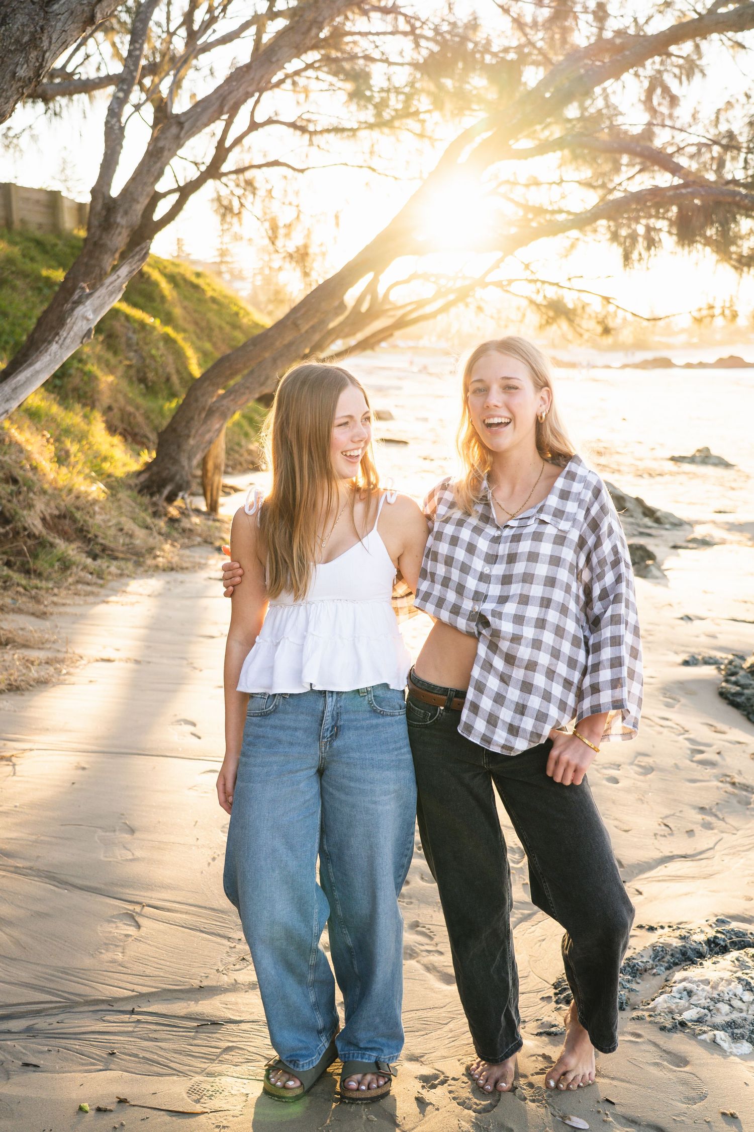 Two teenage girls laughing and posing together on a sandy beach at golden hour sunset.