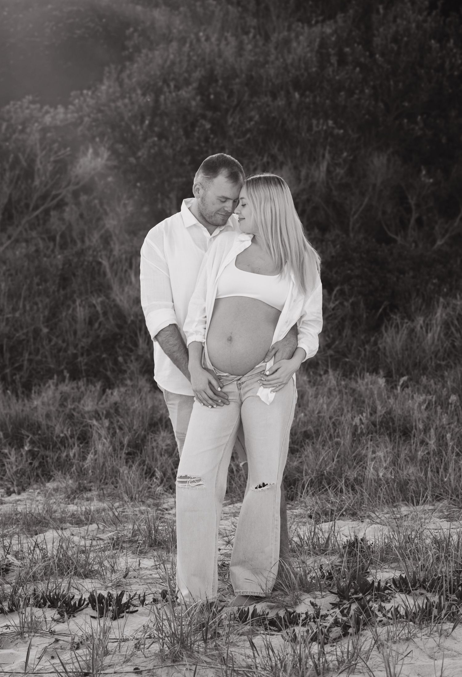 Black and white maternity photo of a couple embracing on a sandy beach, the woman showing her baby bump.