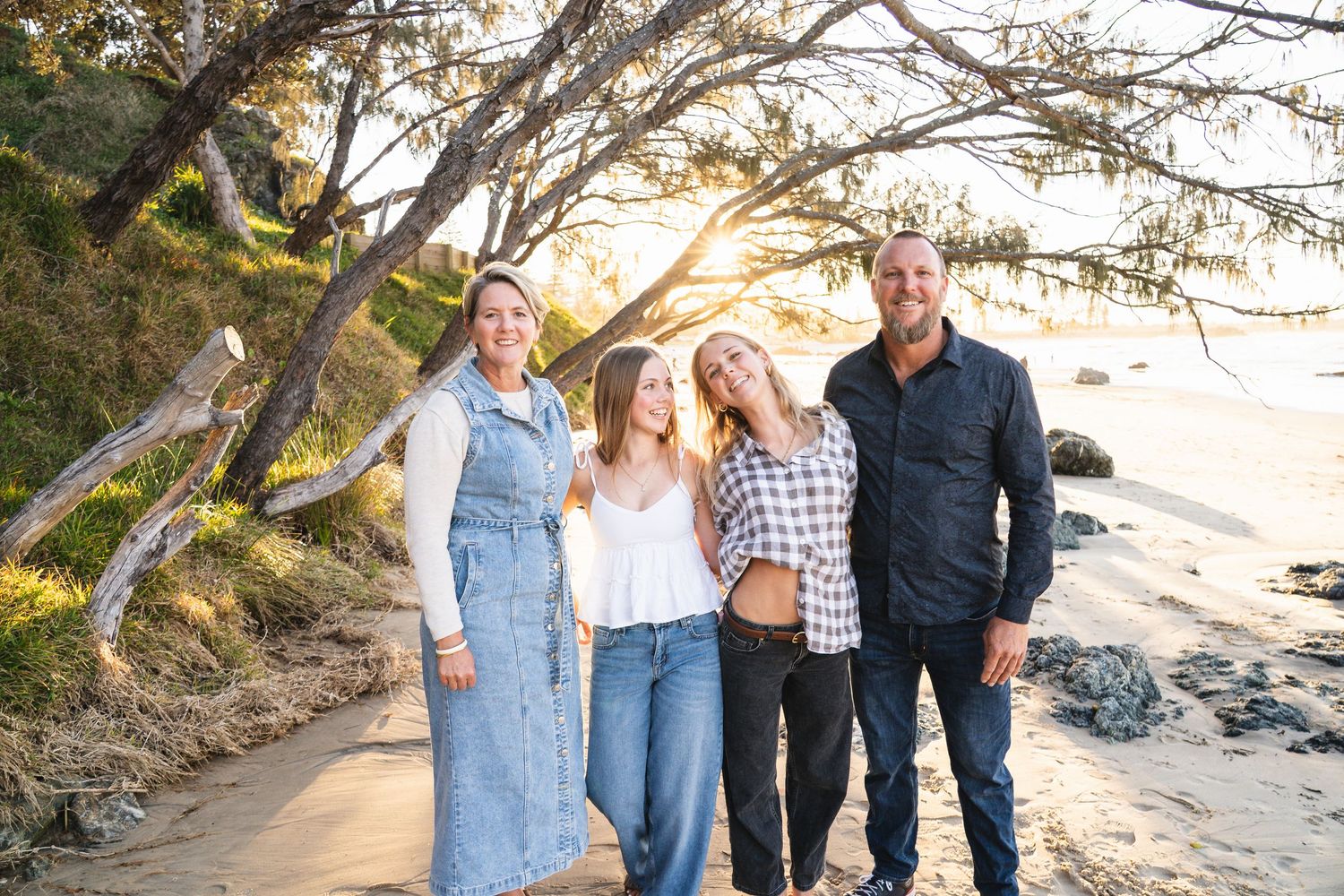 Family of four smiling together on a sandy beach at golden hour with trees and ocean in the background.
