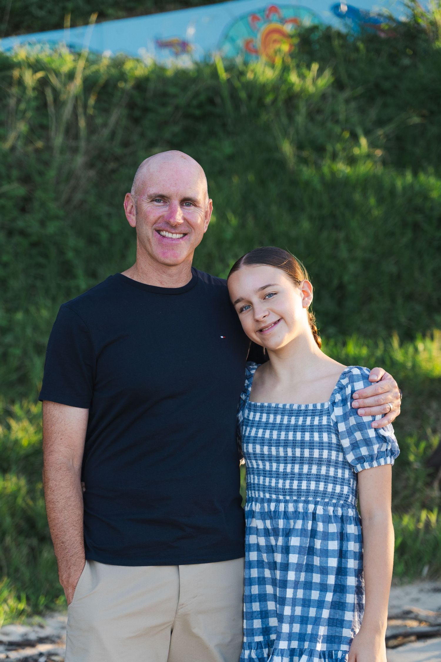 A bald man in a navy t-shirt smiles with a young girl in a blue gingham dress outdoors in natural sunlight.