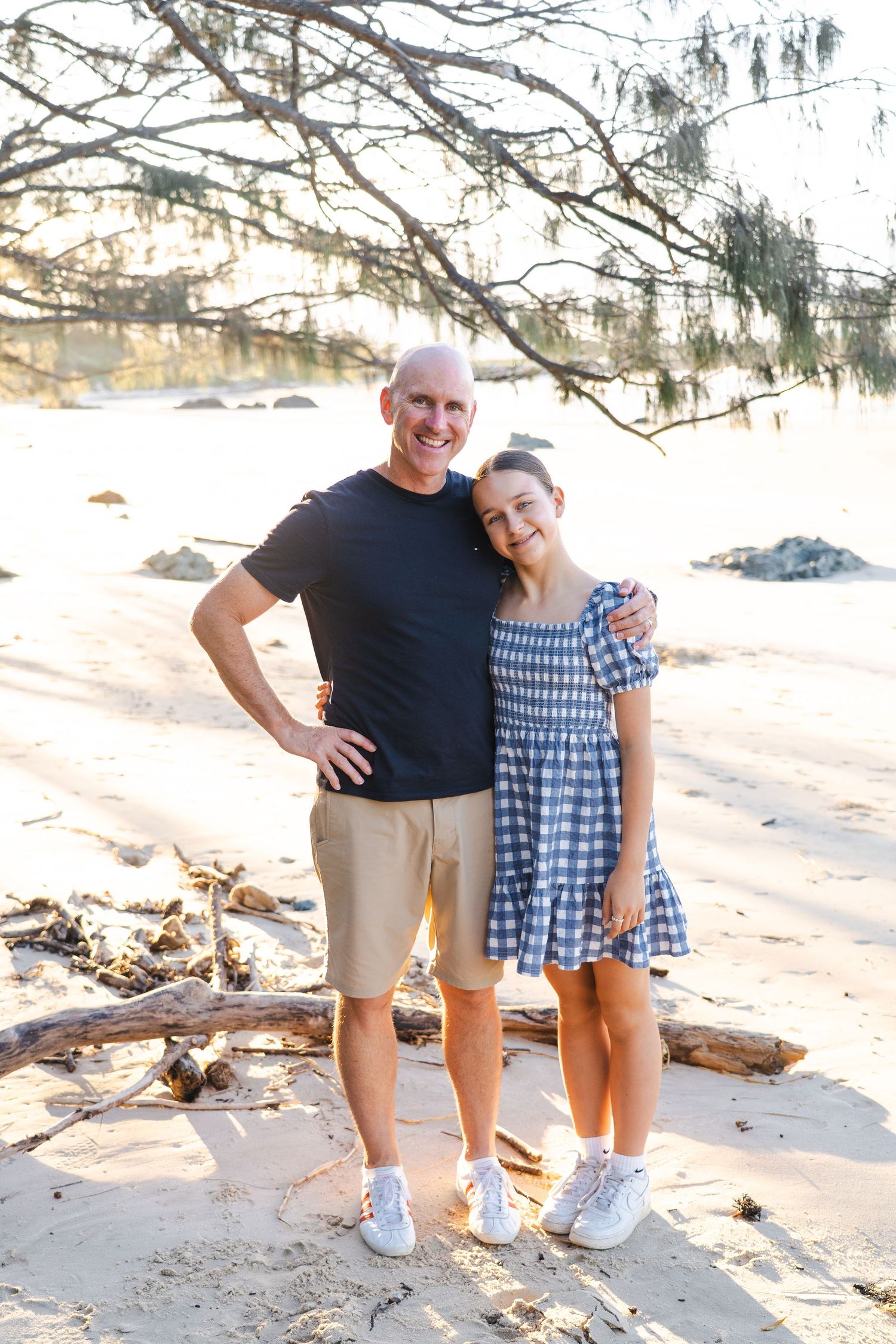 A smiling man and young girl pose together on a sandy beach under tree branches in casual summer outfits.