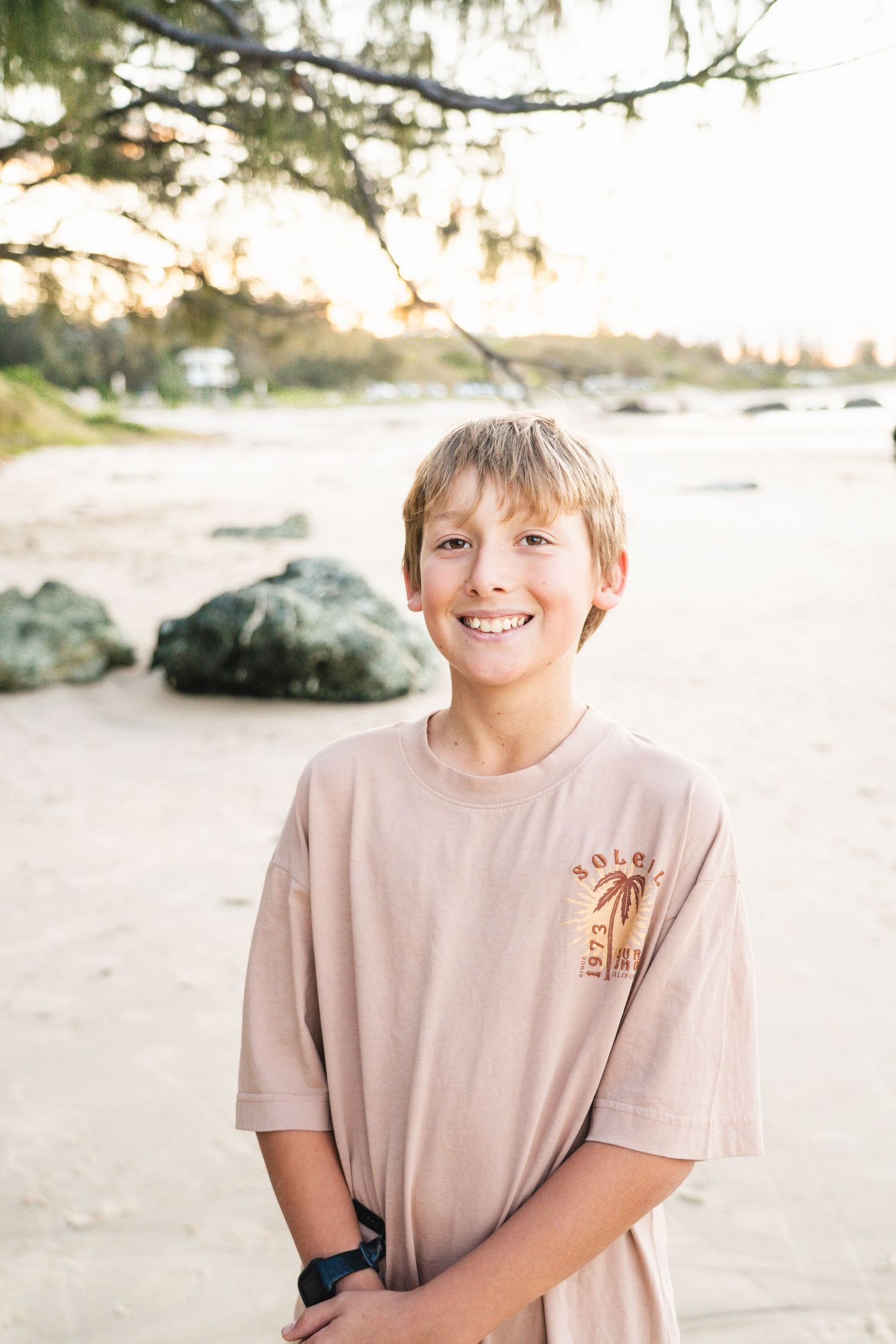 Smiling young boy in a beige graphic t-shirt standing on a sandy beach with rocks and trees in the background.