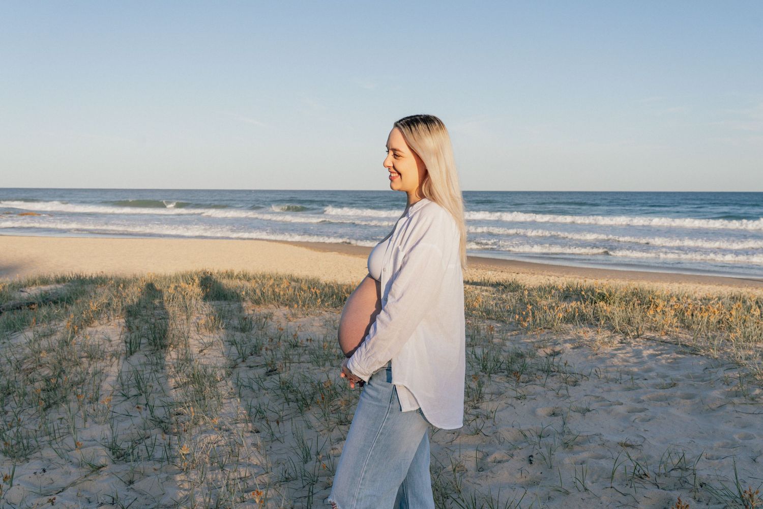 Pregnant woman in white shirt standing on sandy beach with ocean waves and clear blue sky in background.