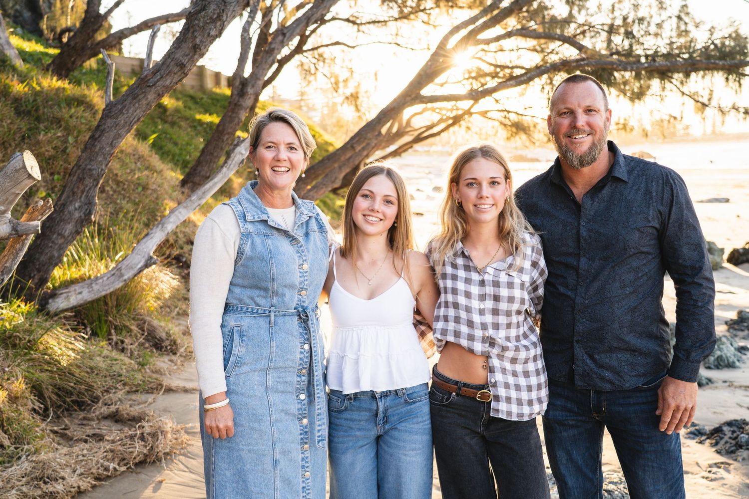 Family of four smiling together on a sunny beach with trees and ocean in the background.