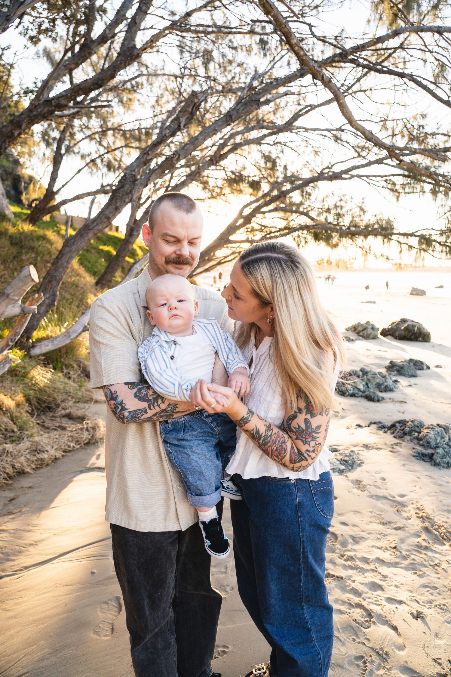 Young couple with tattooed arms lovingly holding their baby boy on a sandy beach path at sunset.