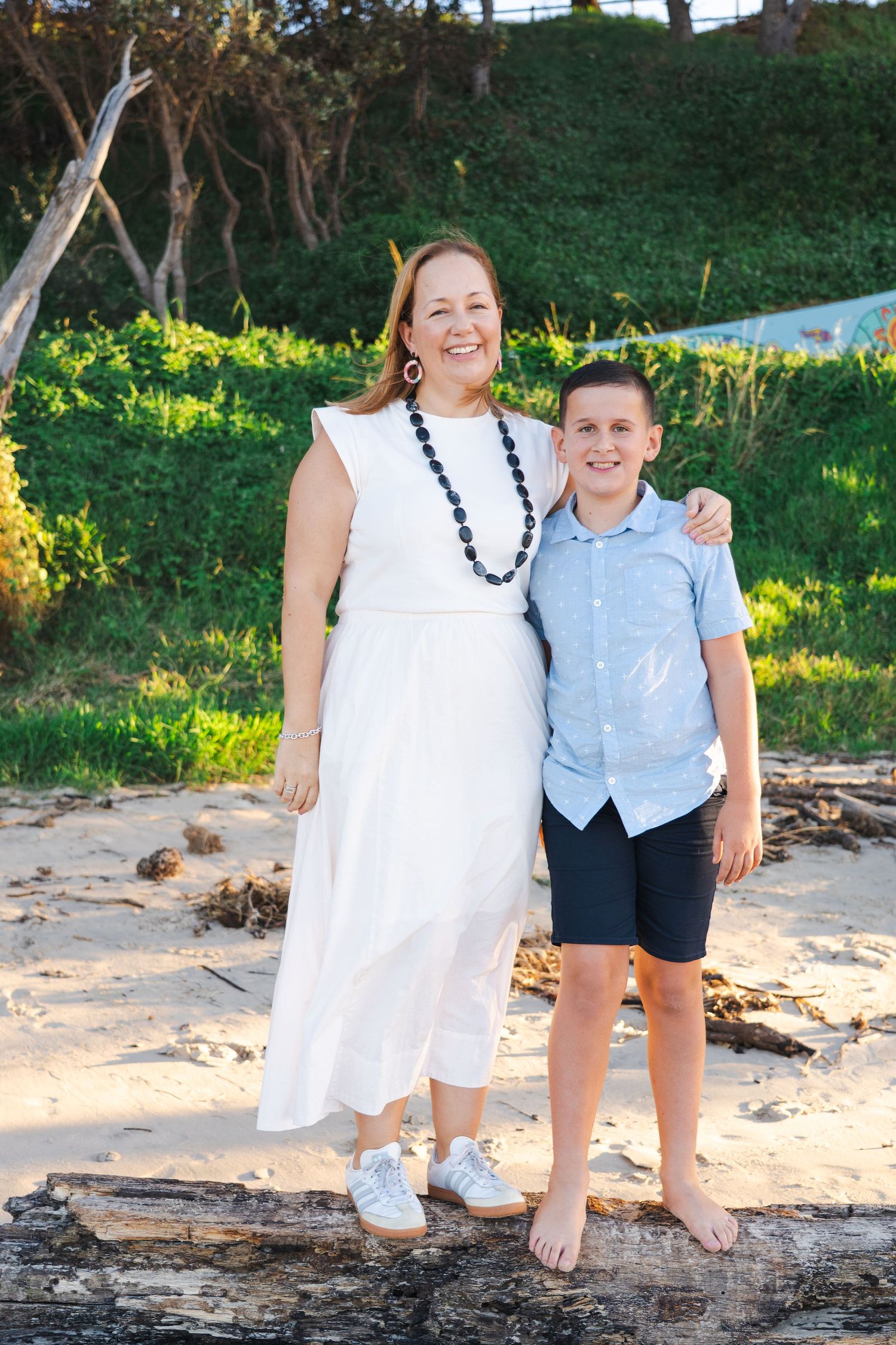 A woman in a white dress and boy in blue shirt smile together on a sandy beach with green trees behind them.