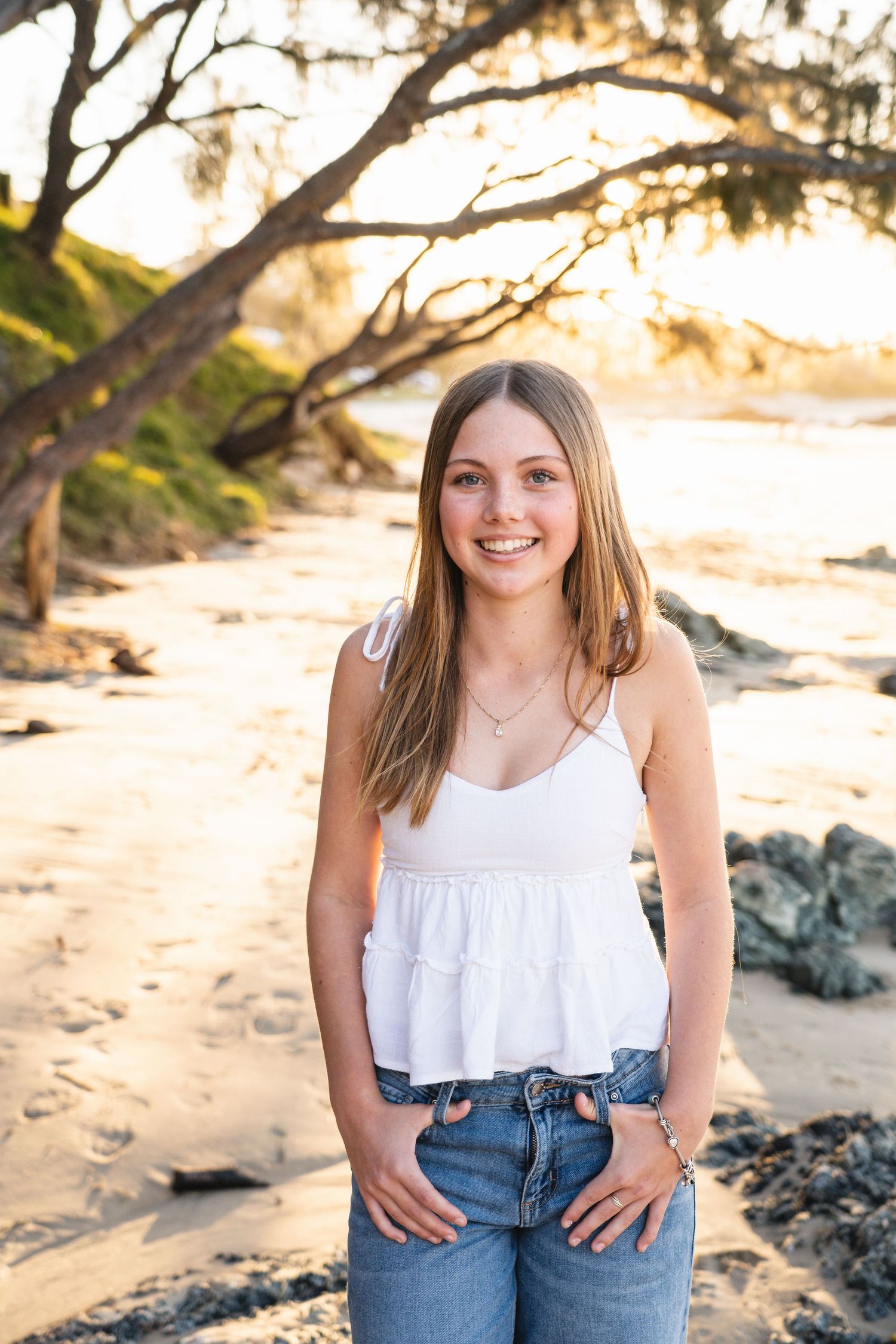 Young woman in white top and jeans smiling on a sandy beach at golden hour with trees and rocks.
