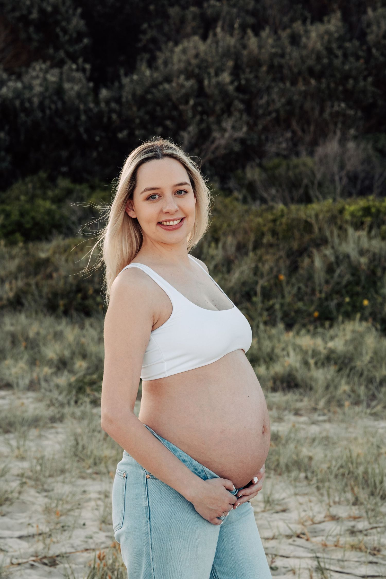 Smiling pregnant woman in white crop top and jeans cradling her baby bump outdoors with greenery background.