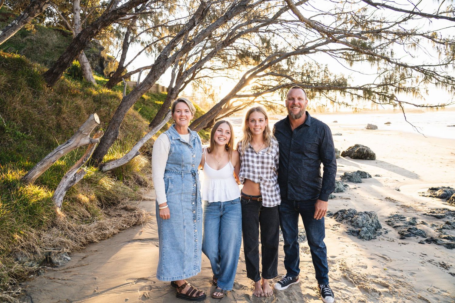 Family of four smiling together on a sandy beach path surrounded by trees at golden hour.