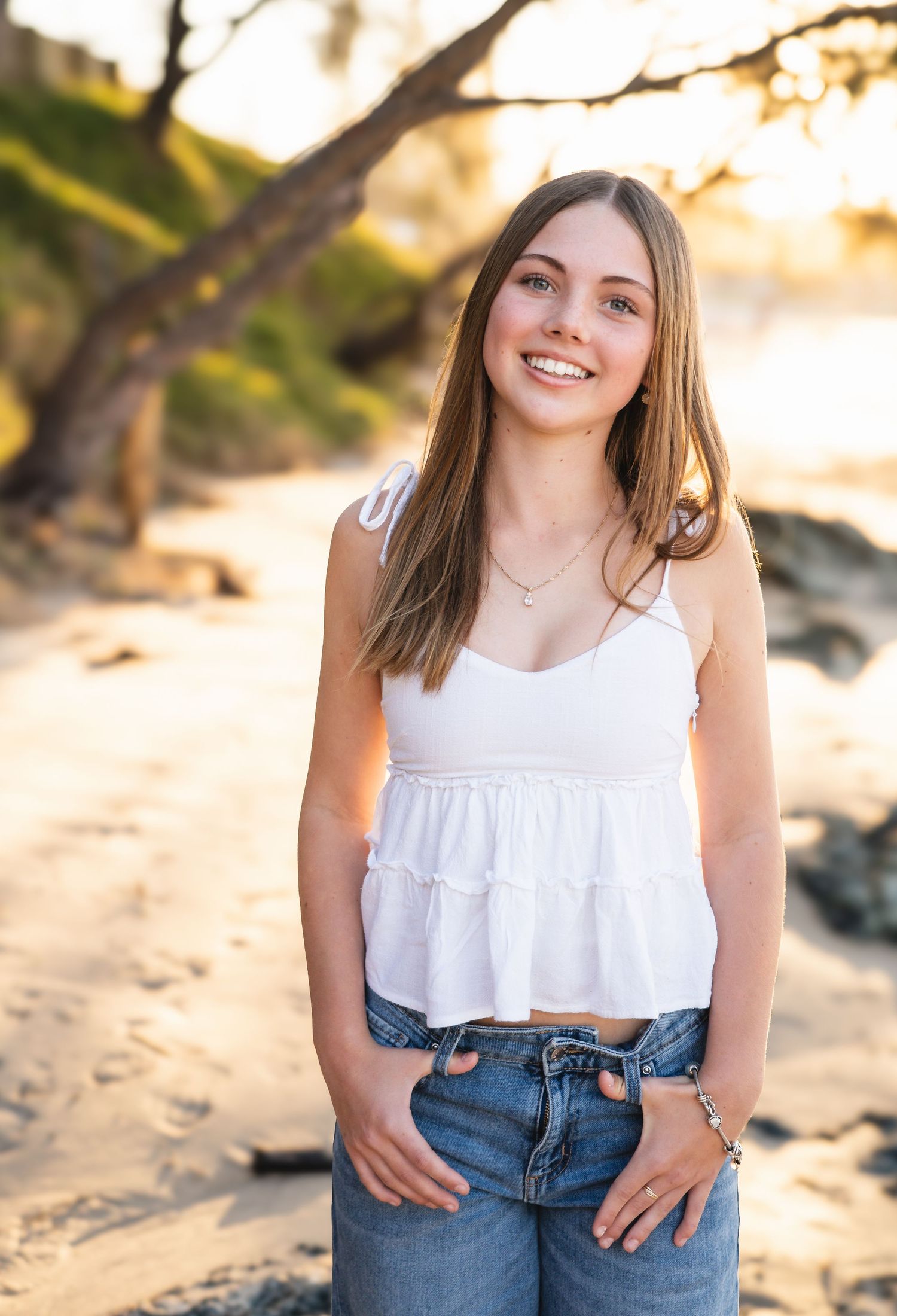 Smiling young woman in white crop top and jeans poses on a sandy beach at golden hour.