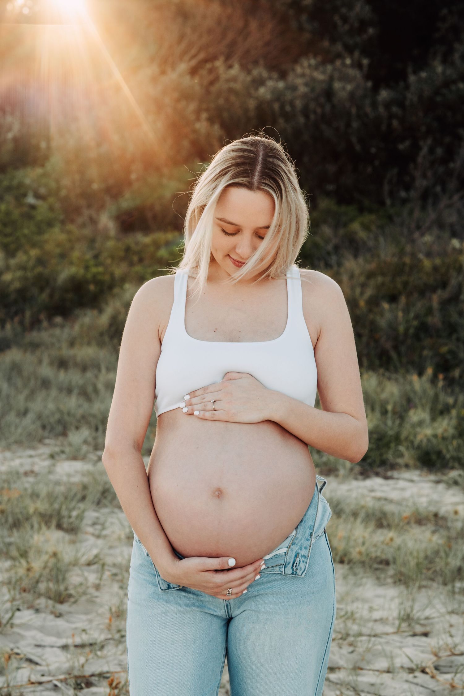 Pregnant woman in white crop top and jeans cradling her baby bump outdoors in golden sunset light.