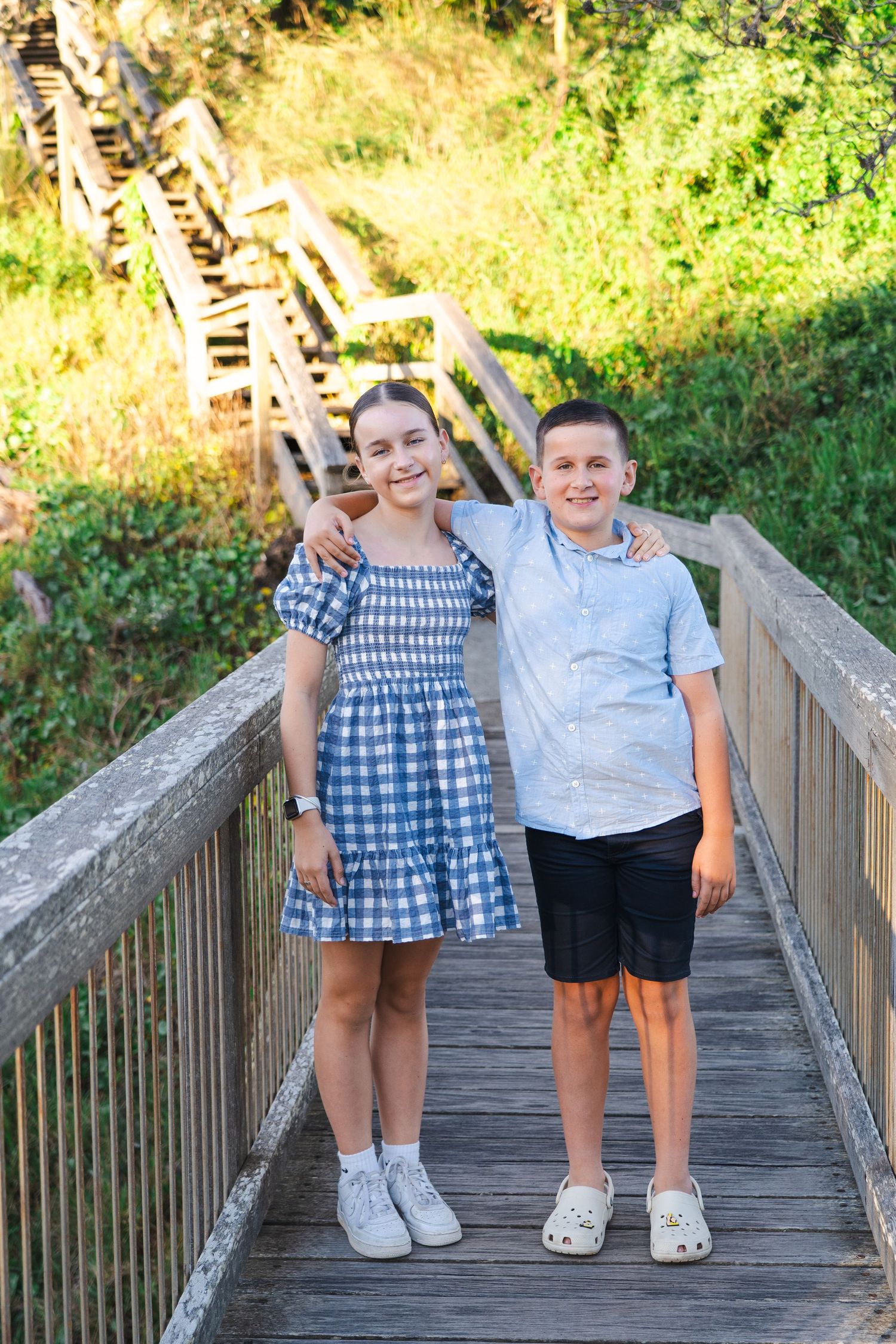 Two smiling kids pose on a wooden boardwalk bridge surrounded by lush greenery on a sunny day.