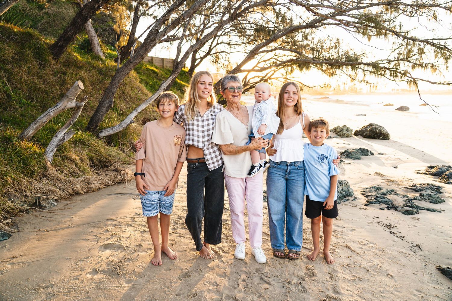 Family of six posing together on a sandy beach at golden hour with rocky coastline and trees in background.