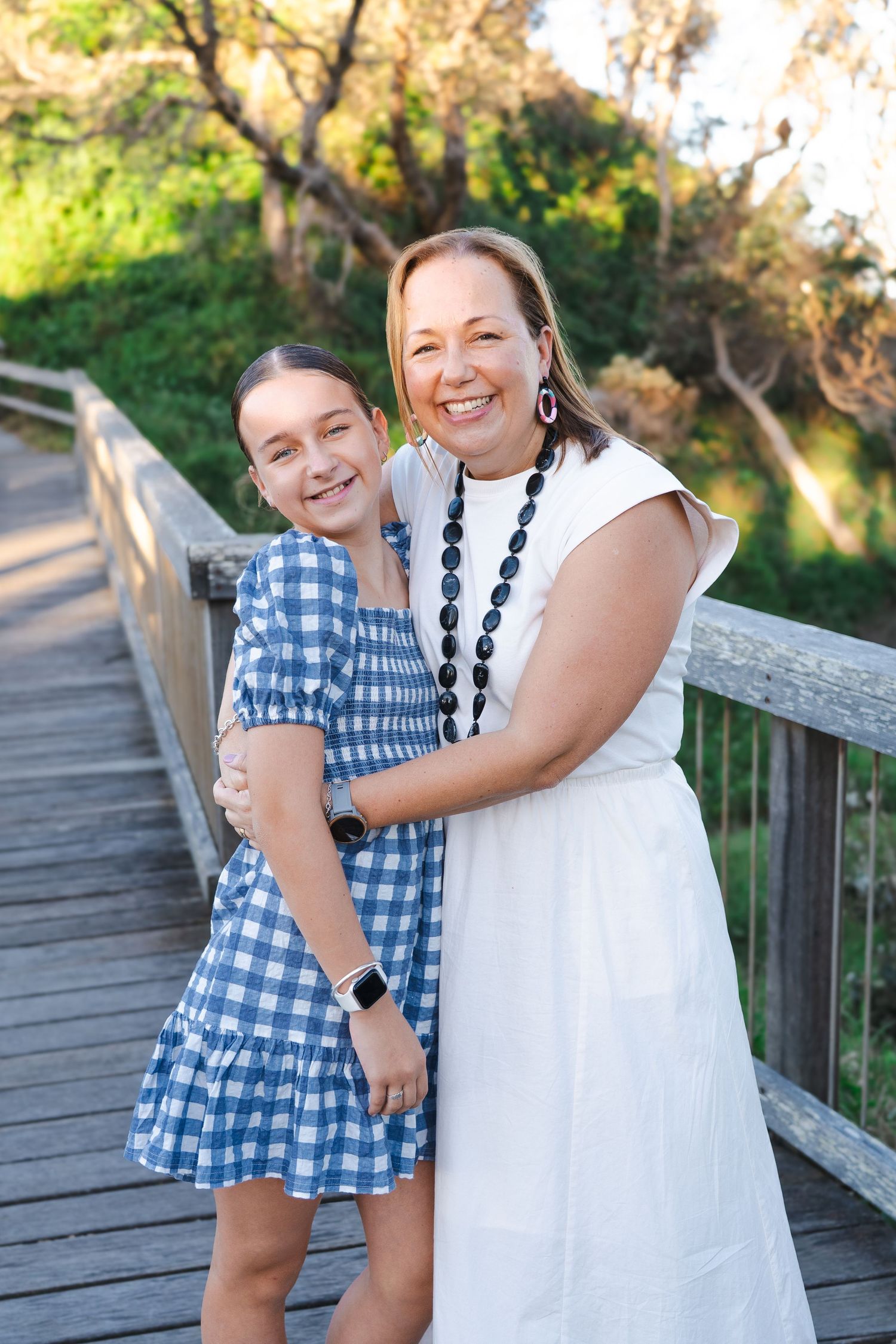 A woman and young girl in matching smiles embrace on a wooden bridge surrounded by greenery.