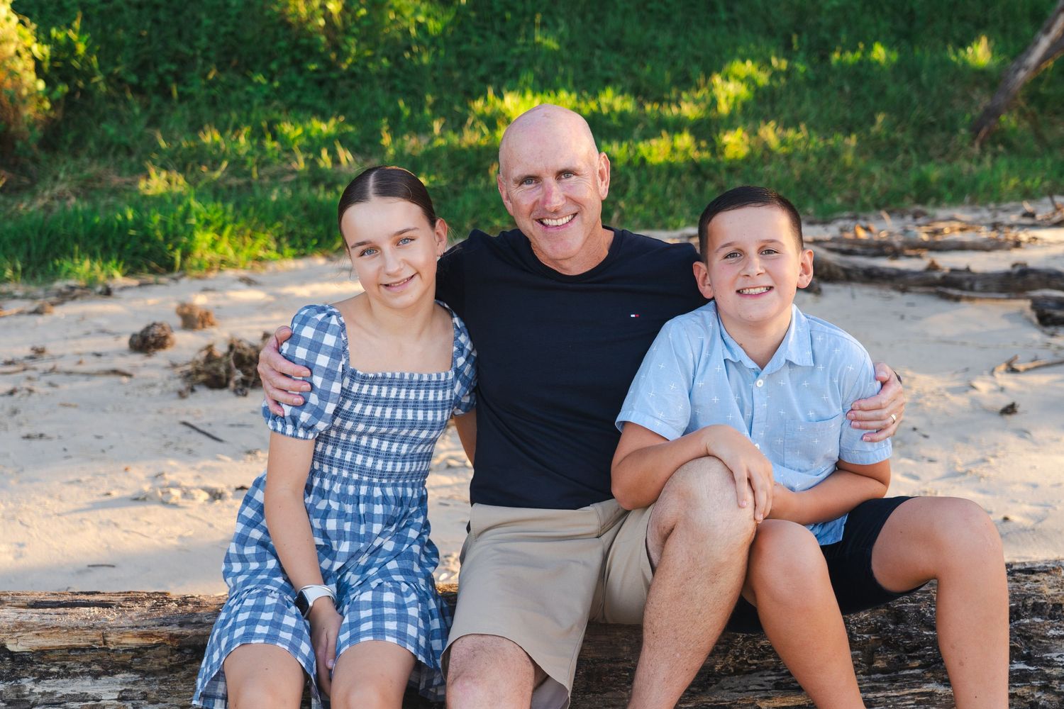 A bald man smiles with two children on a sandy beach surrounded by green trees in warm sunlight.