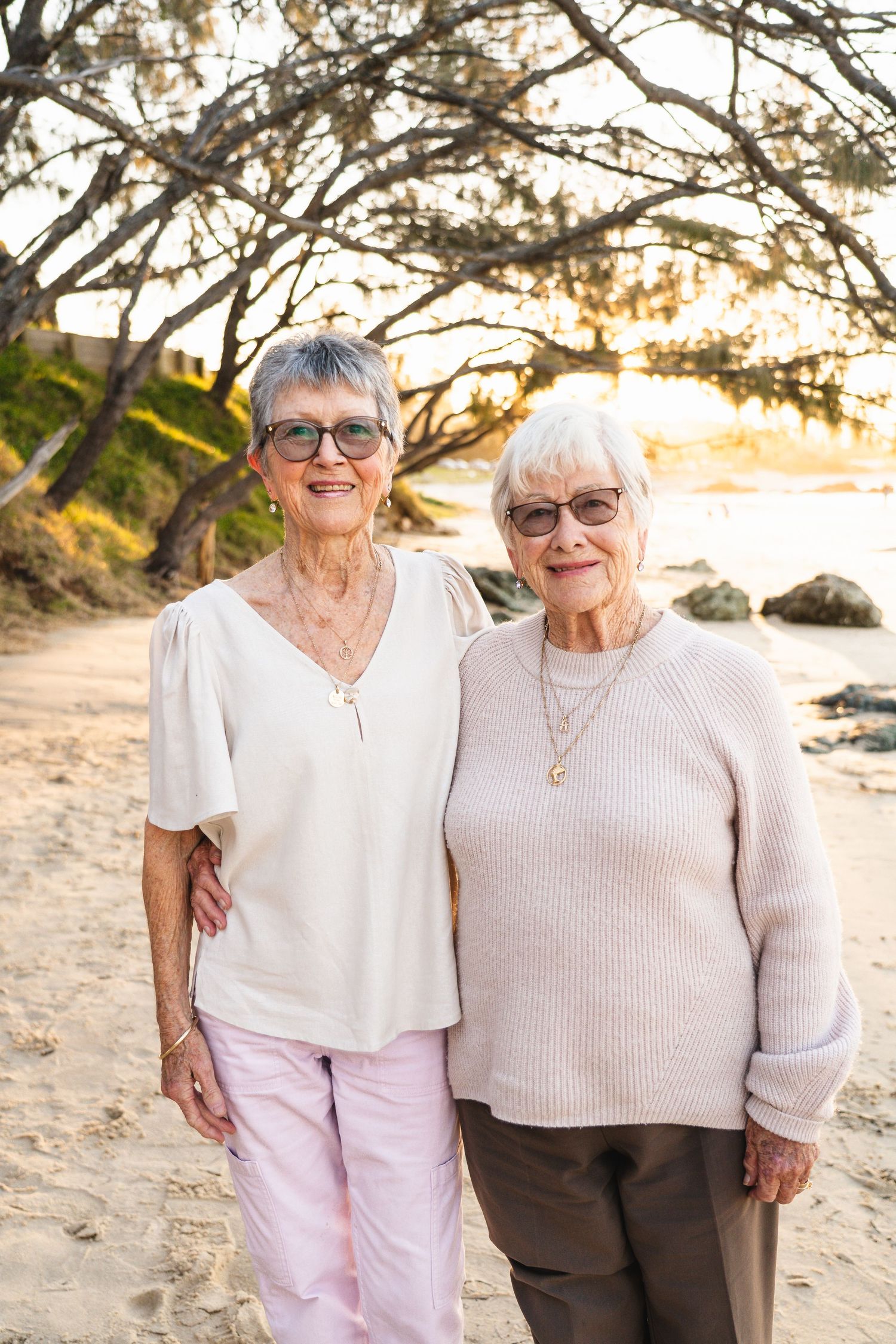 Two elderly women smiling together on a sandy beach at sunset, wearing casual pastel clothing and glasses.