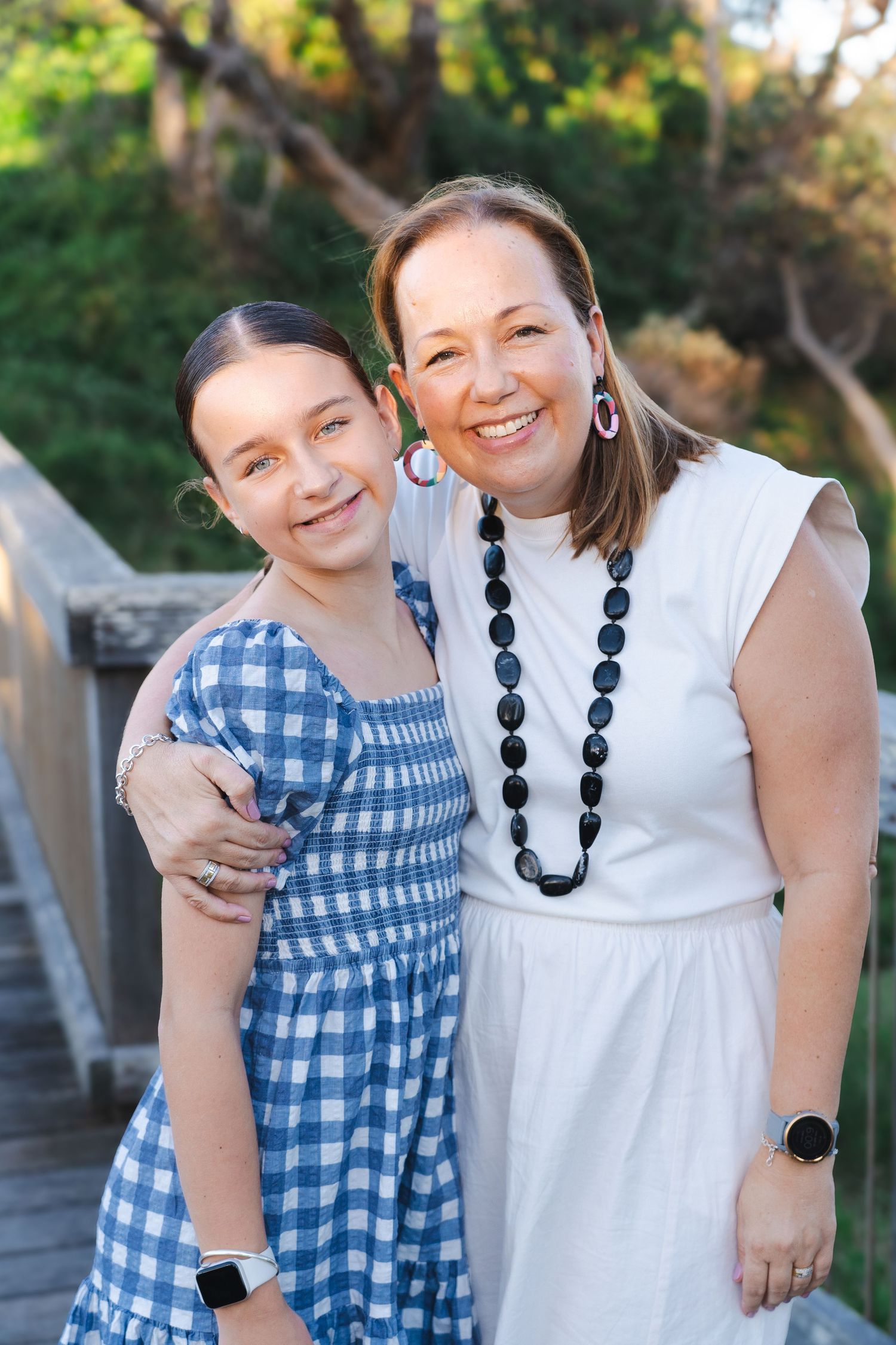 A woman and young girl in blue gingham dress smile together on a wooden bridge surrounded by green trees.