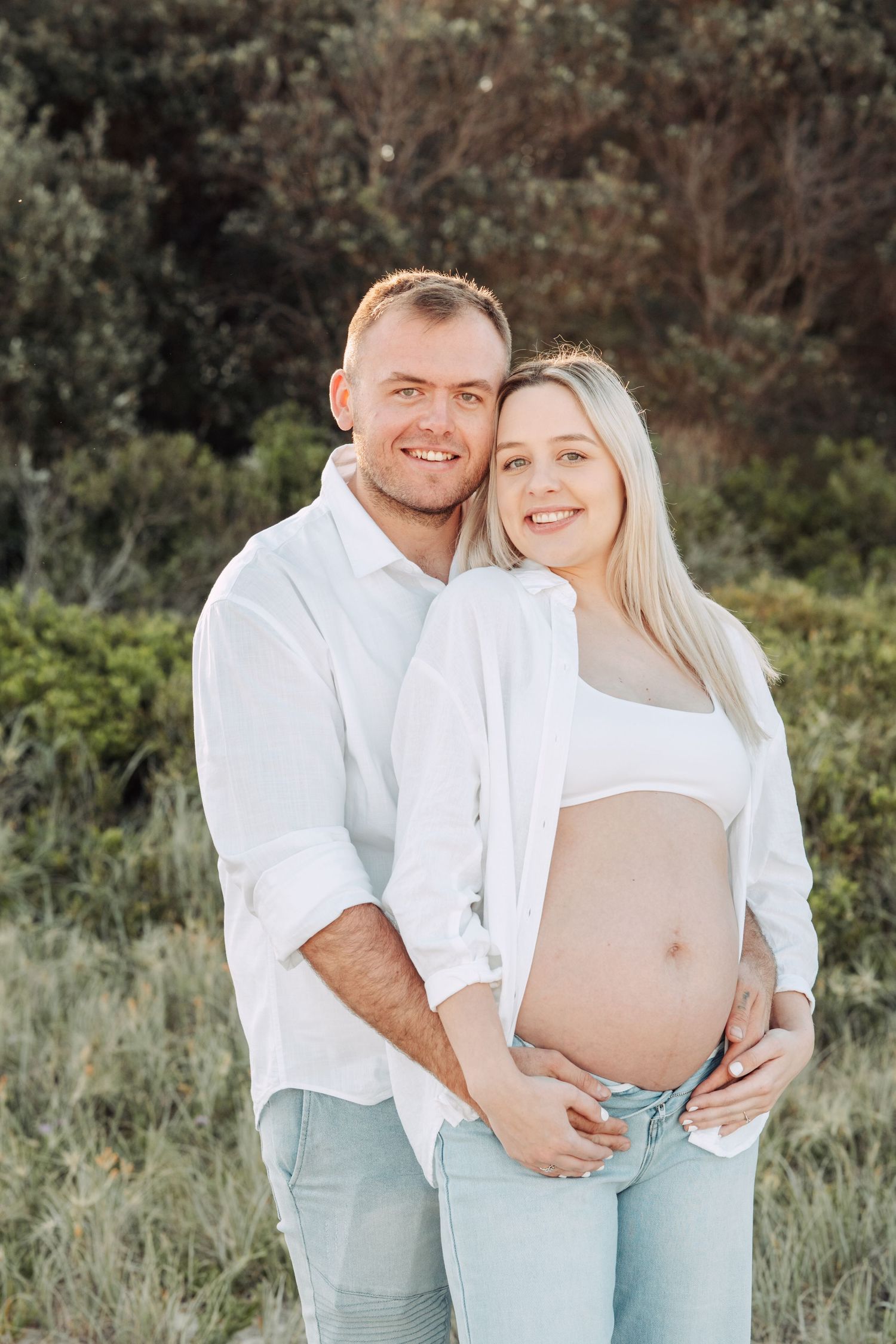 A smiling couple outdoors, man in white shirt embracing pregnant woman showing baby bump, surrounded by green shrubs.
