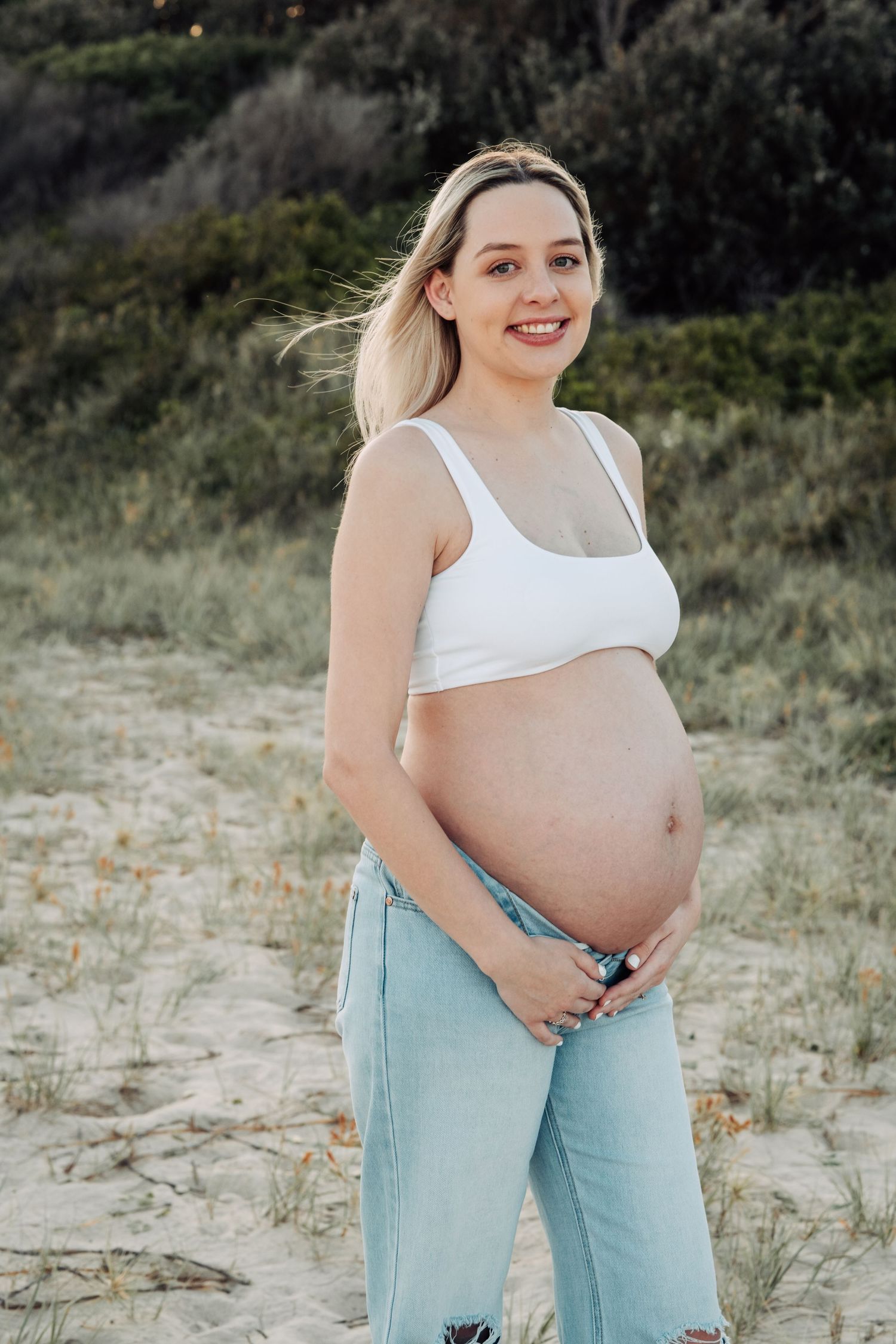 Smiling pregnant blonde woman in white crop top and jeans holding baby bump on sandy beach with greenery behind her.