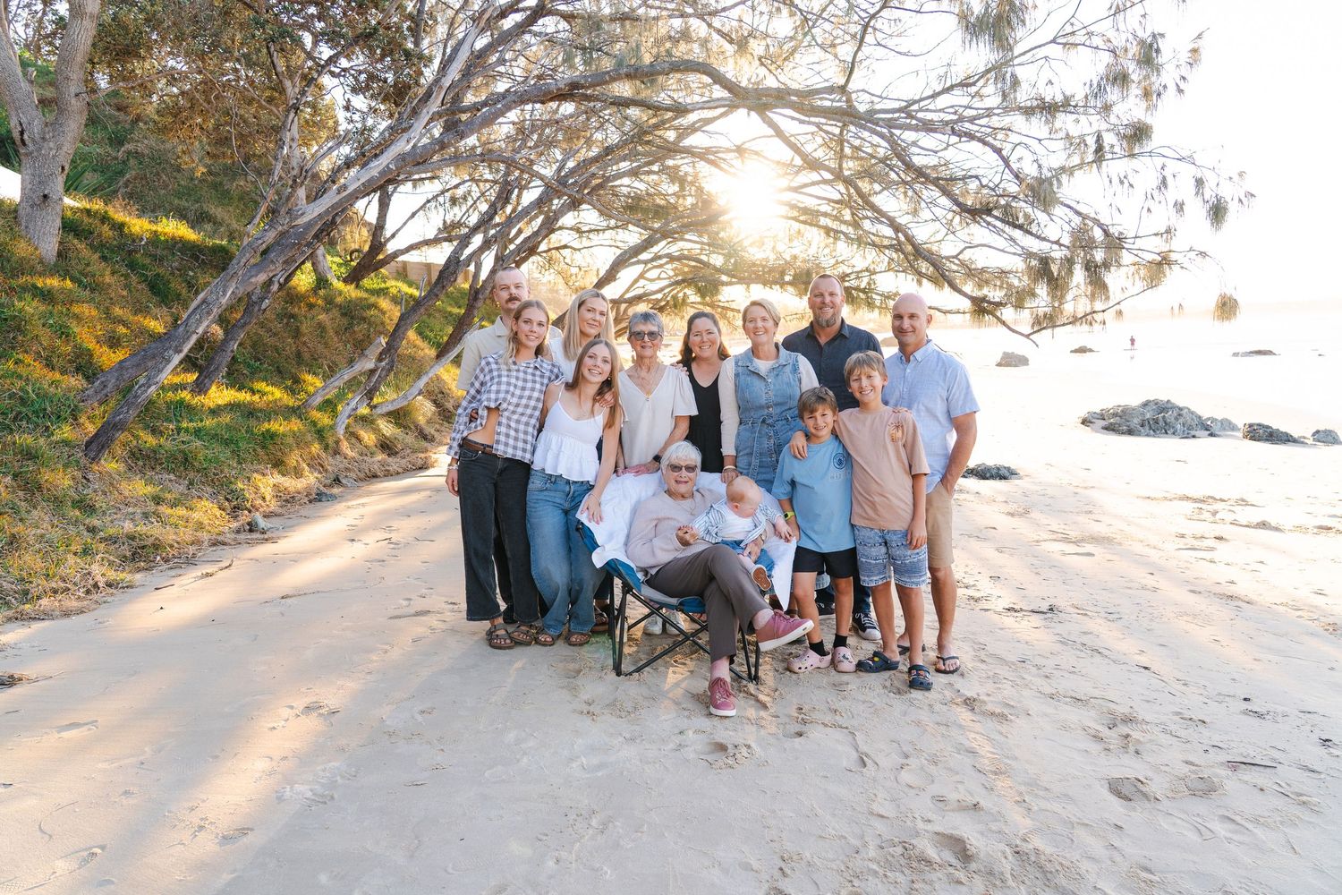 Family group photo on a sandy beach at golden hour, posing under trees with ocean in the background.
