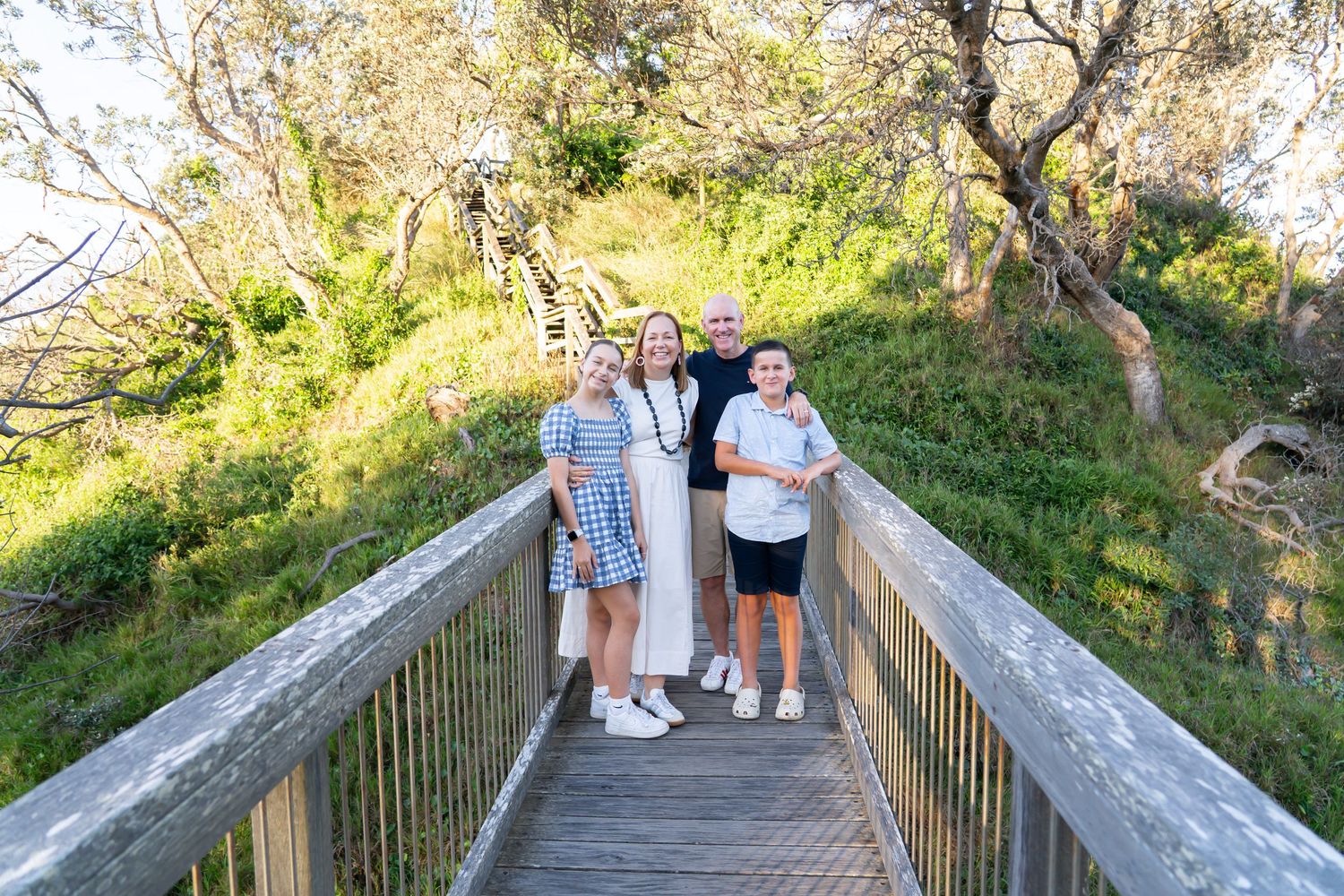 Family of four smiling on a wooden bridge surrounded by lush green trees and nature trail.