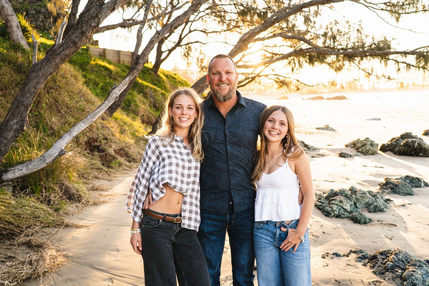 A man and two young women smile together on a sandy beach path surrounded by trees at golden hour.