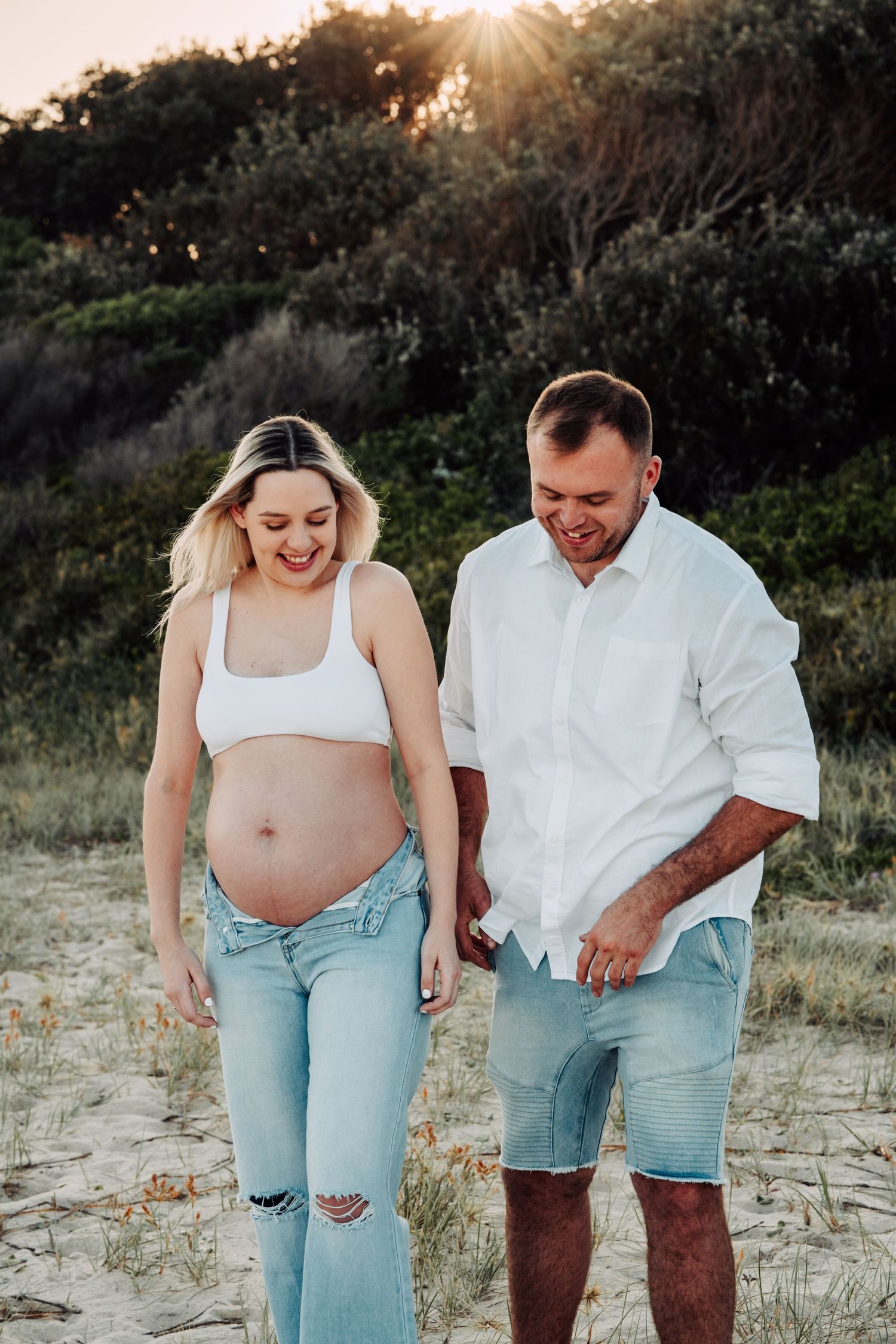 Pregnant couple walking on sandy beach at sunset, woman in white crop top and ripped jeans holding hands with man in white shirt.