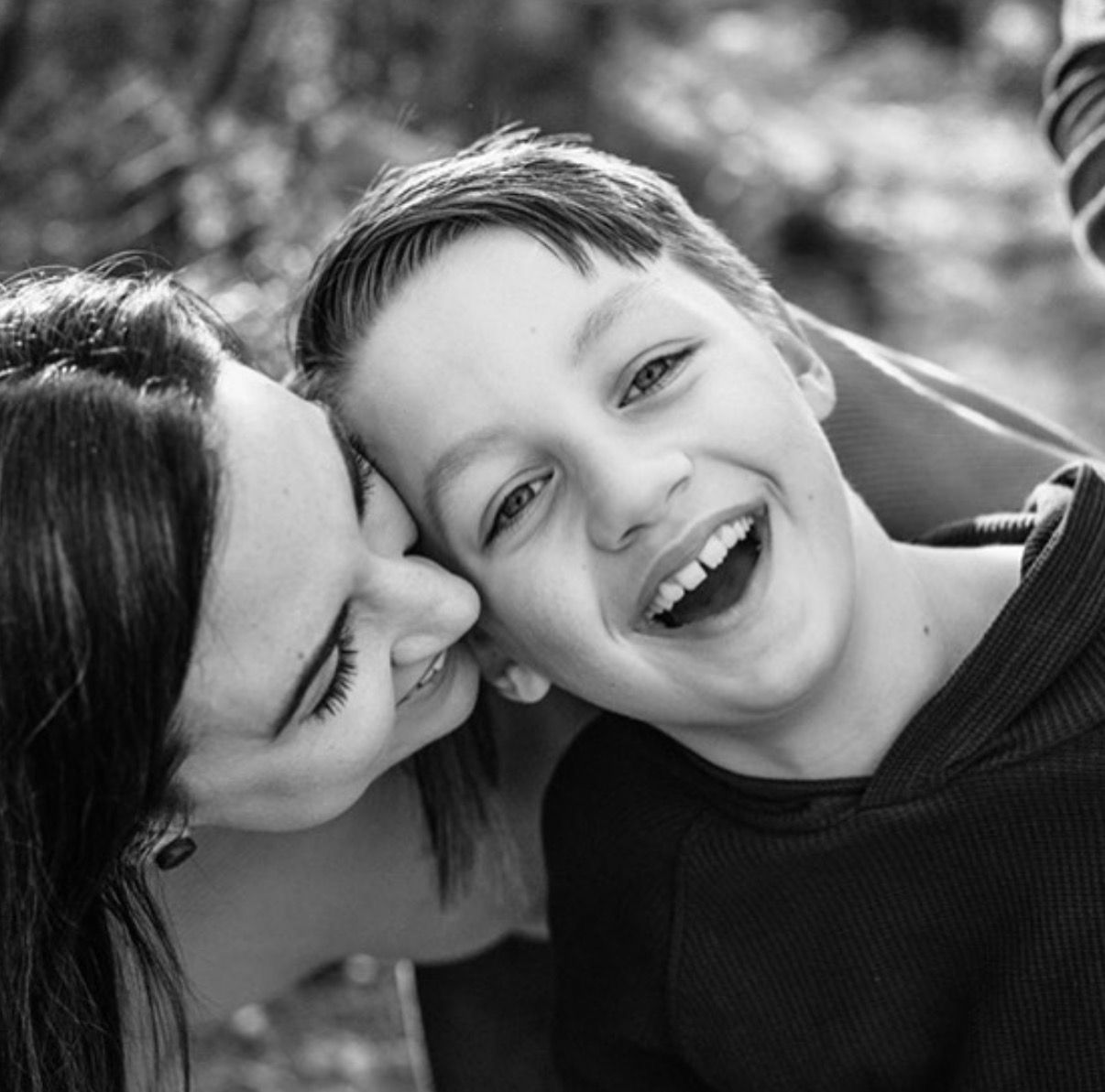 Black and white photo of a woman kissing a laughing boy on the cheek outdoors.