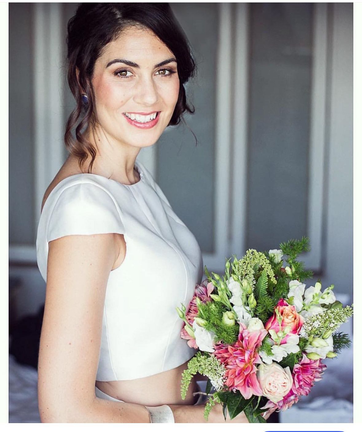 A bride in a white dress holding a vibrant pink and coral bouquet of flowers smiles in a bright indoor setting.