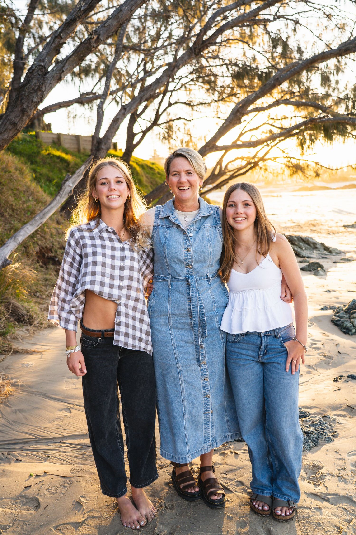 Three women smiling together on a sandy beach at golden hour, dressed in casual summer outfits near coastal trees.