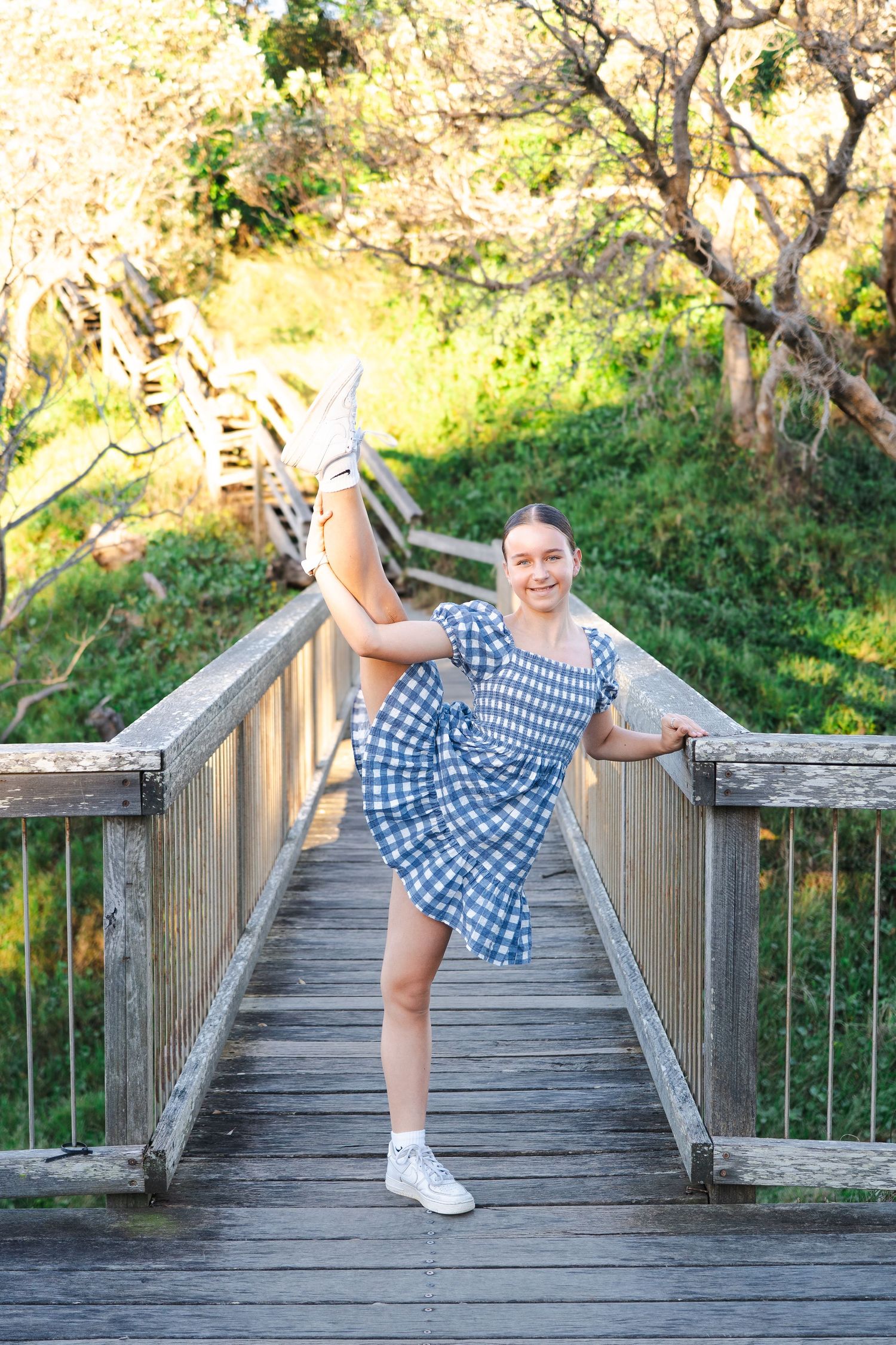 Smiling young woman in blue gingham dress doing a high kick on a wooden bridge surrounded by green trees.