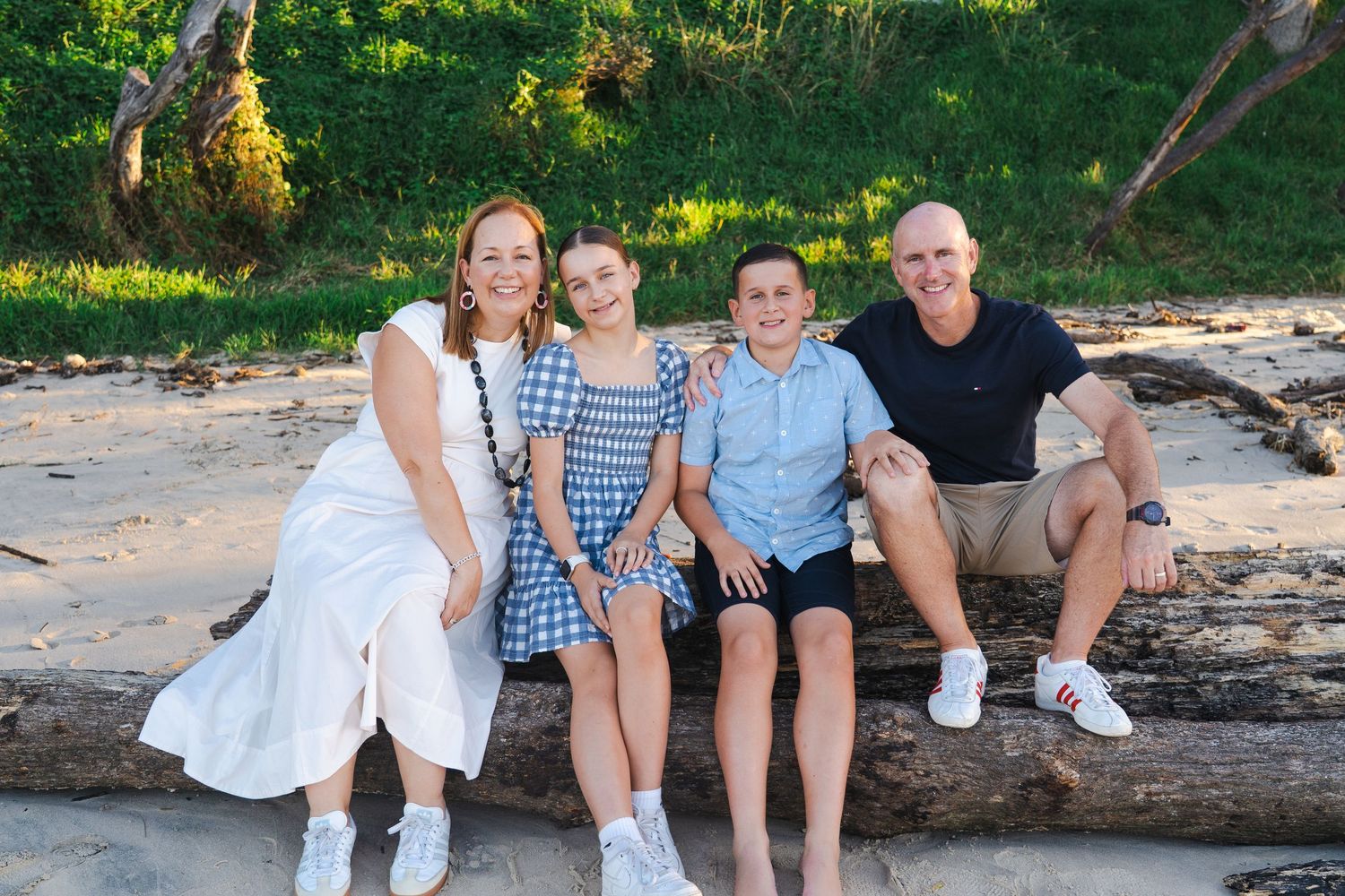 Family of four smiling together on a driftwood log at a tropical beach, wearing casual summer outfits.