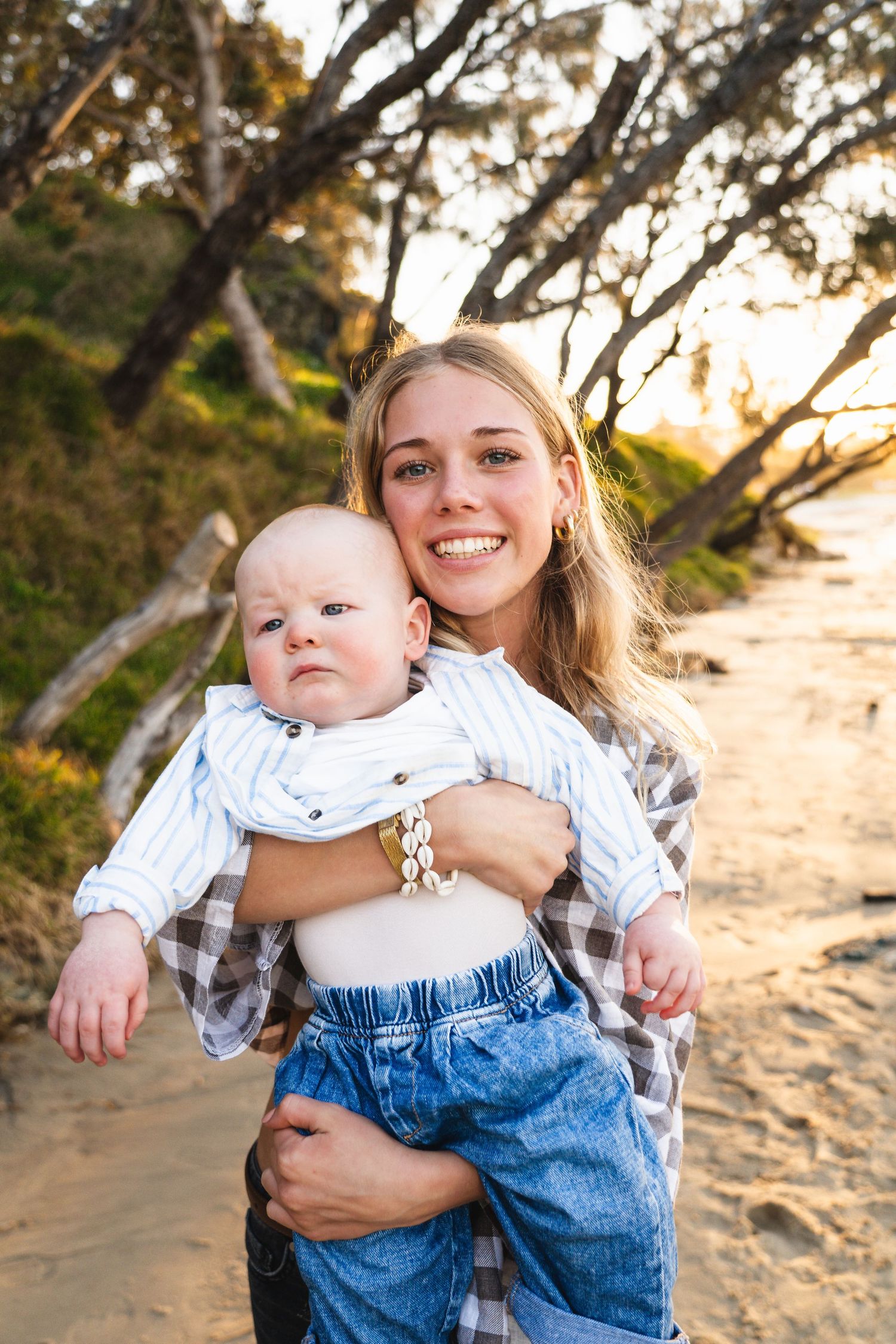 Young smiling blonde woman holding a baby on a sandy beach at golden hour with trees in the background.