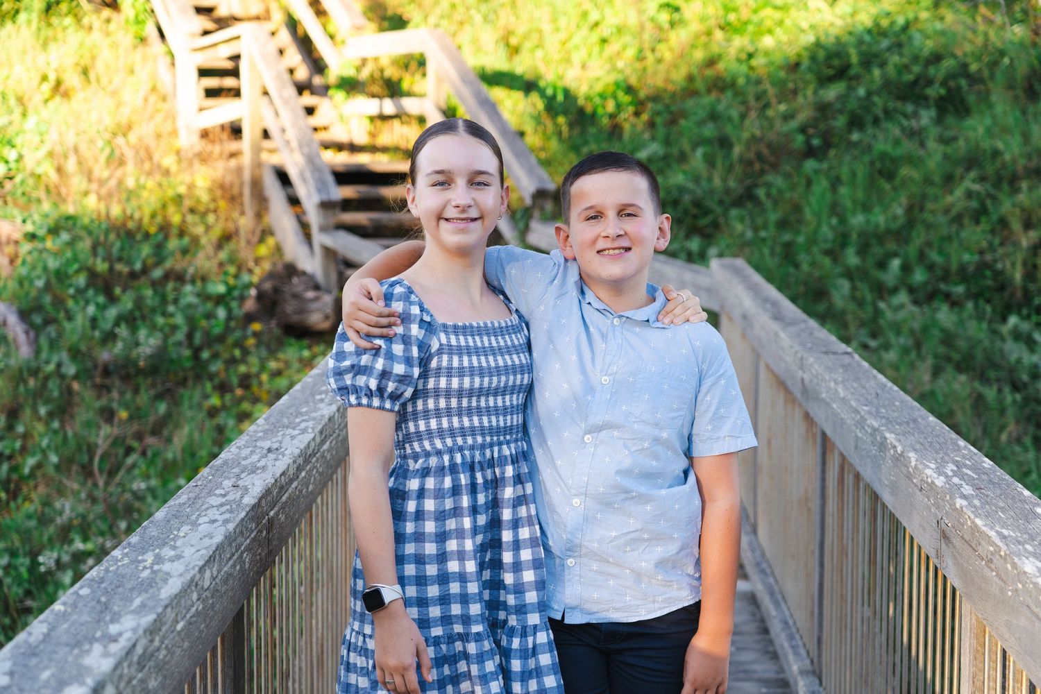 Two smiling children pose on a wooden bridge surrounded by lush greenery on a sunny day.