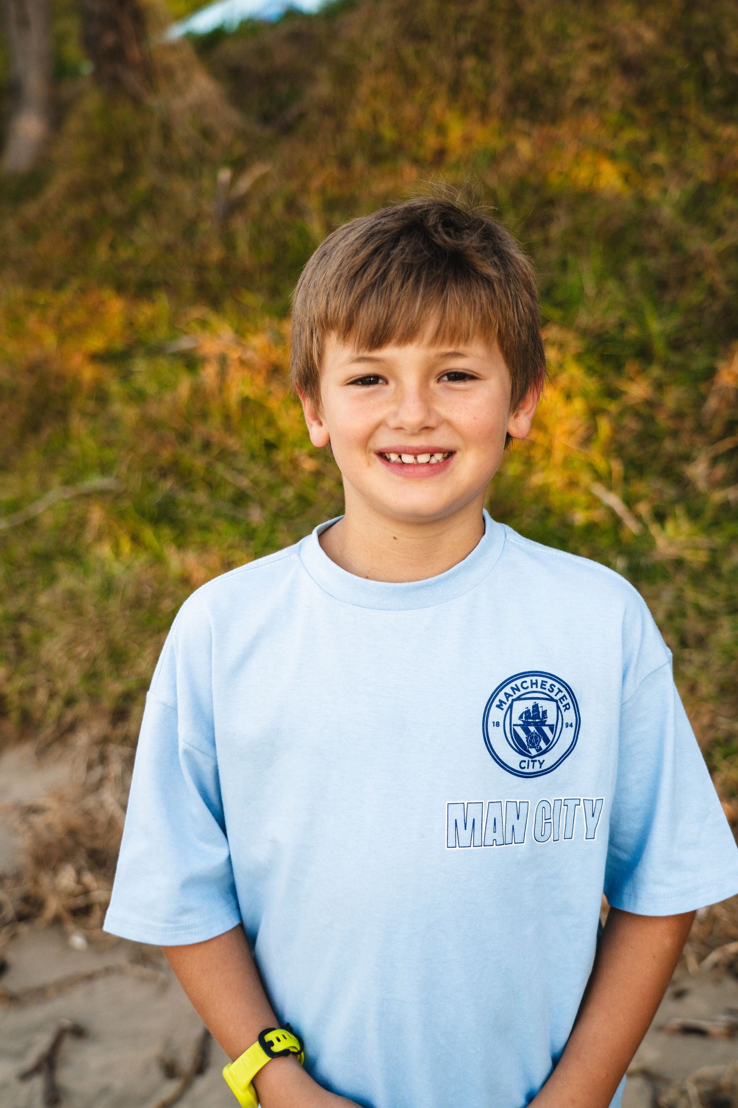 Smiling boy in a light blue Manchester City t-shirt posing outdoors with green and yellow foliage in the background.