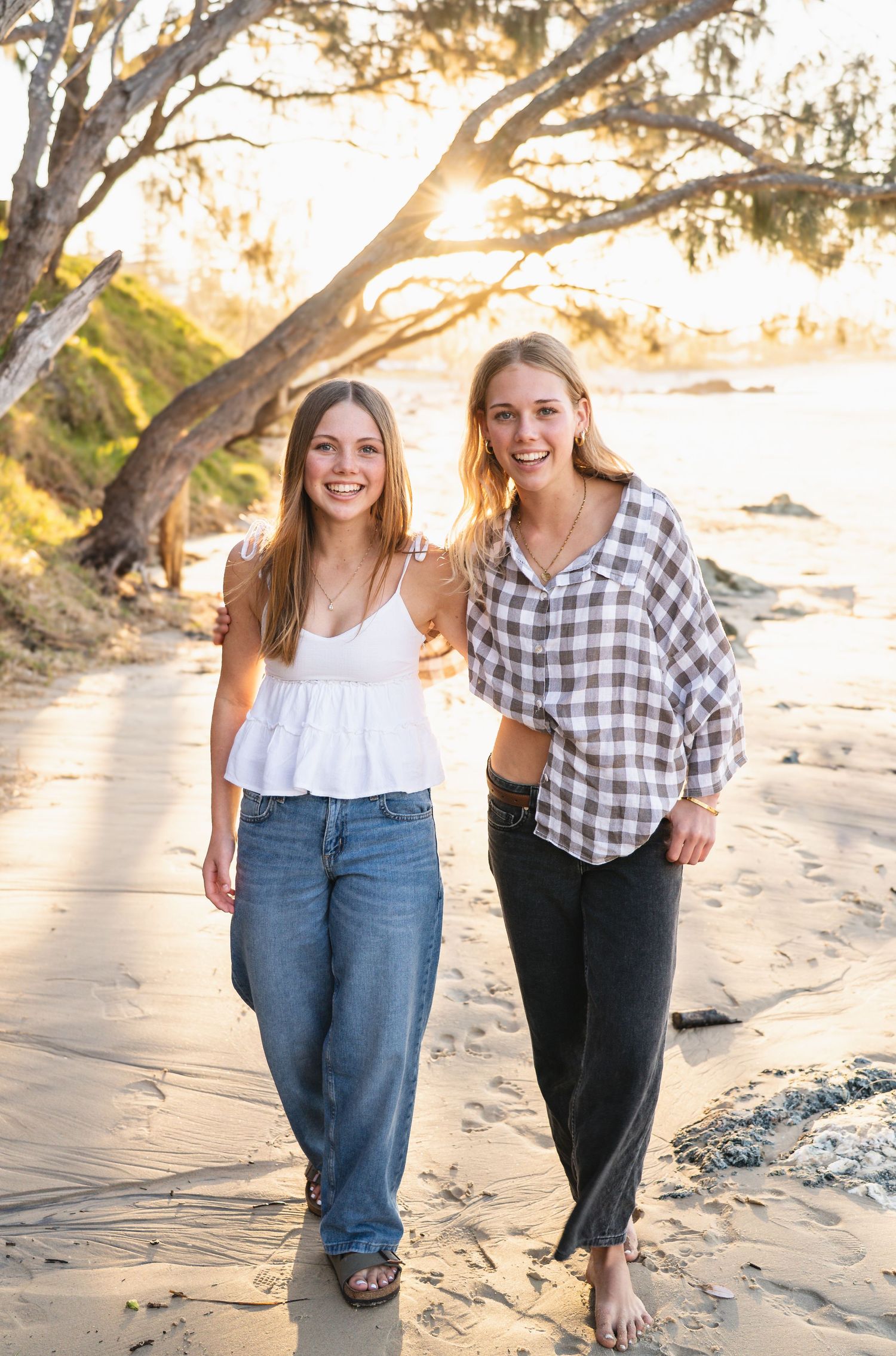 Two smiling young women walk along a sandy beach at golden sunset, one in white top and jeans, other in plaid shirt.