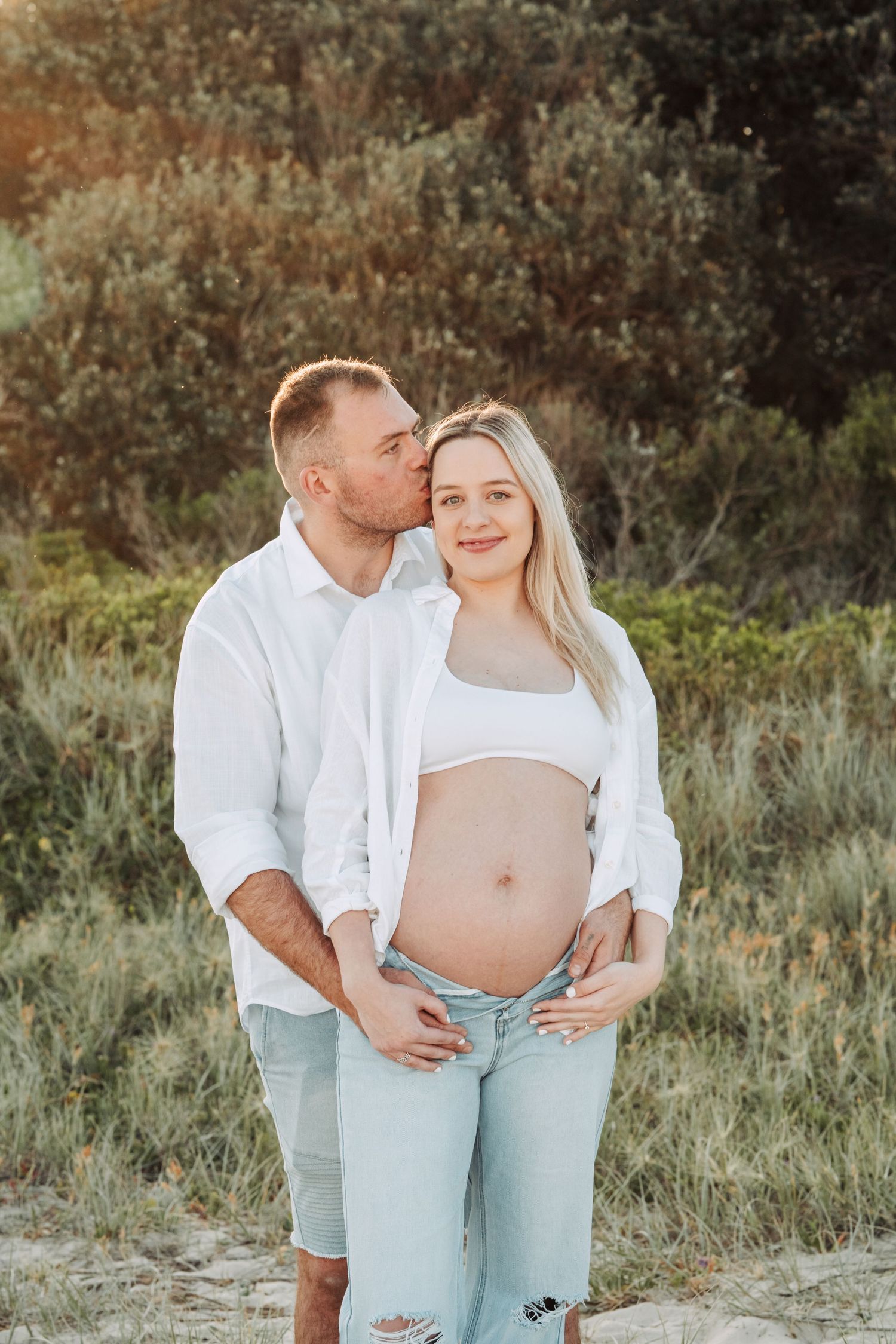 Couple outdoors at golden hour, man kissing pregnant woman's cheek while cradling her baby bump, wearing white tops and jeans.