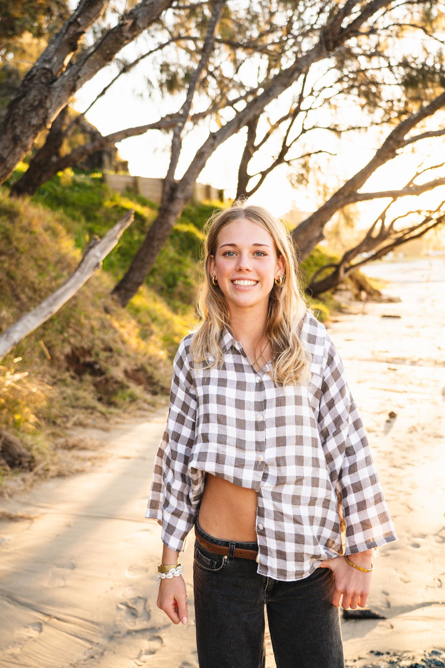 Smiling blonde woman in plaid shirt and jeans standing on sandy beach path surrounded by trees at golden hour.