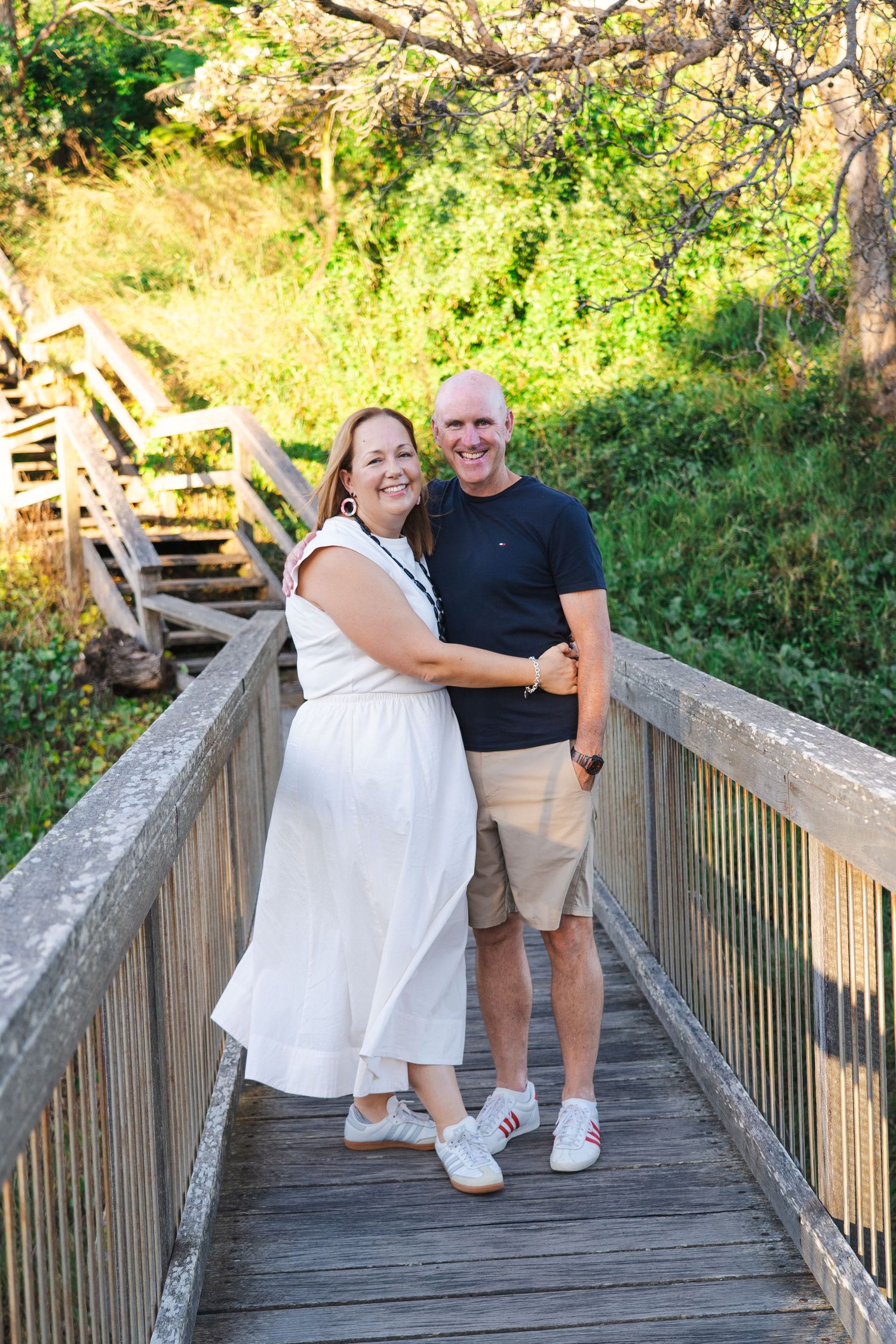 A smiling couple embraces on a wooden bridge surrounded by lush greenery, woman in white dress, man in navy shirt.