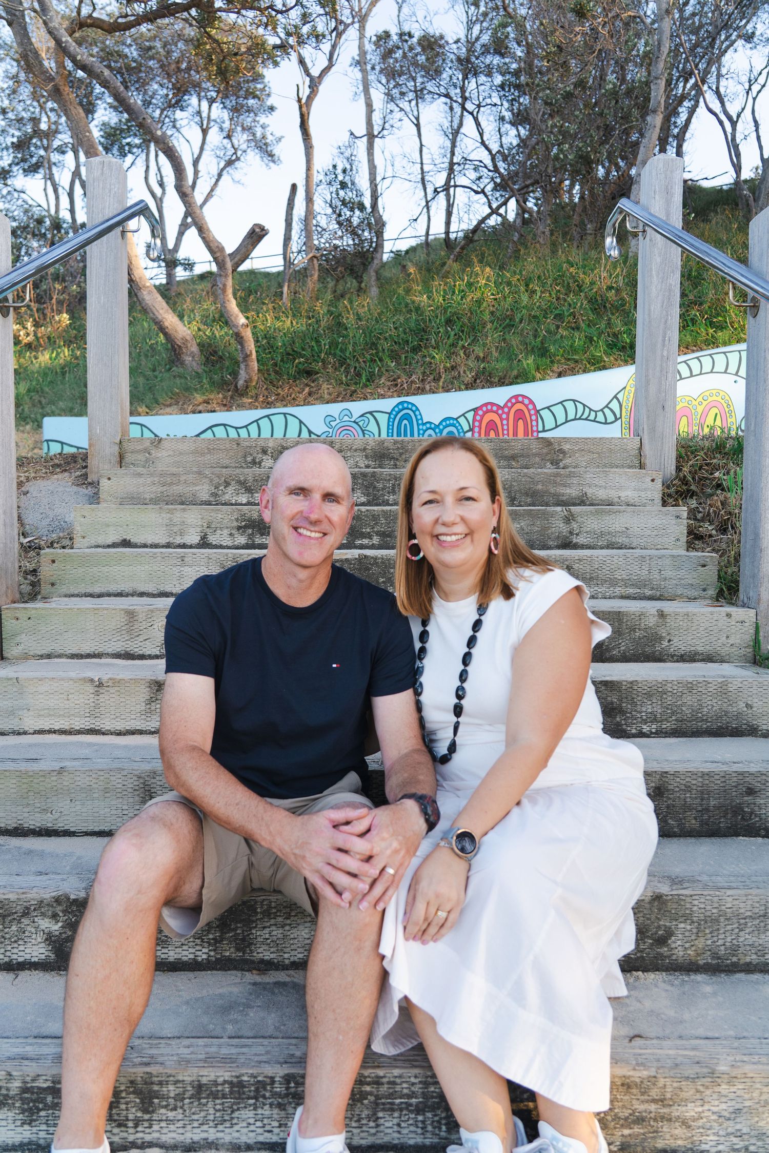 A smiling couple sits together on painted outdoor stairs surrounded by trees in a casual outdoor setting.