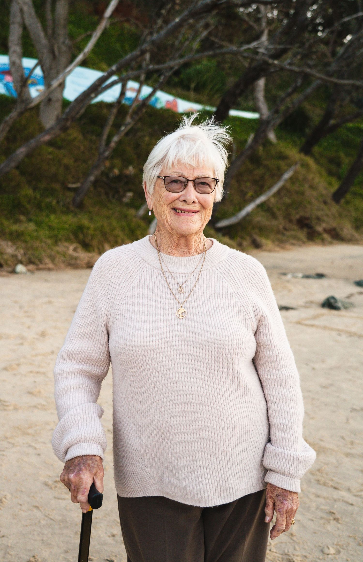 Elderly woman with white hair and glasses smiling on a beach holding a walking cane.