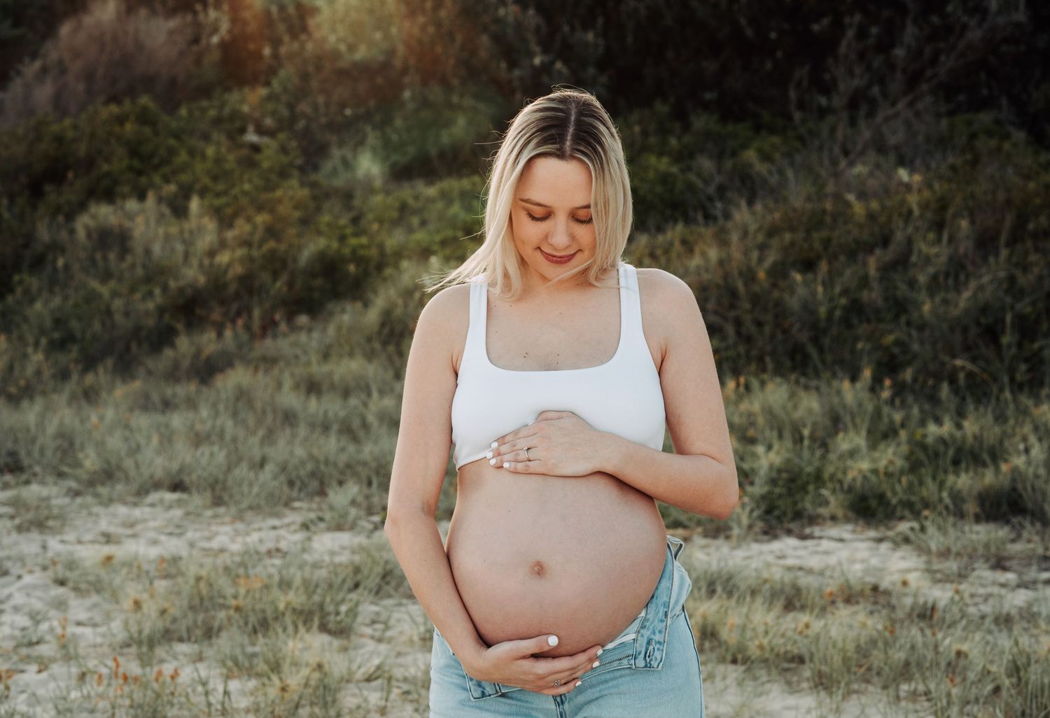 Pregnant woman in white crop top and jeans cradling her baby bump outdoors in a natural field setting.