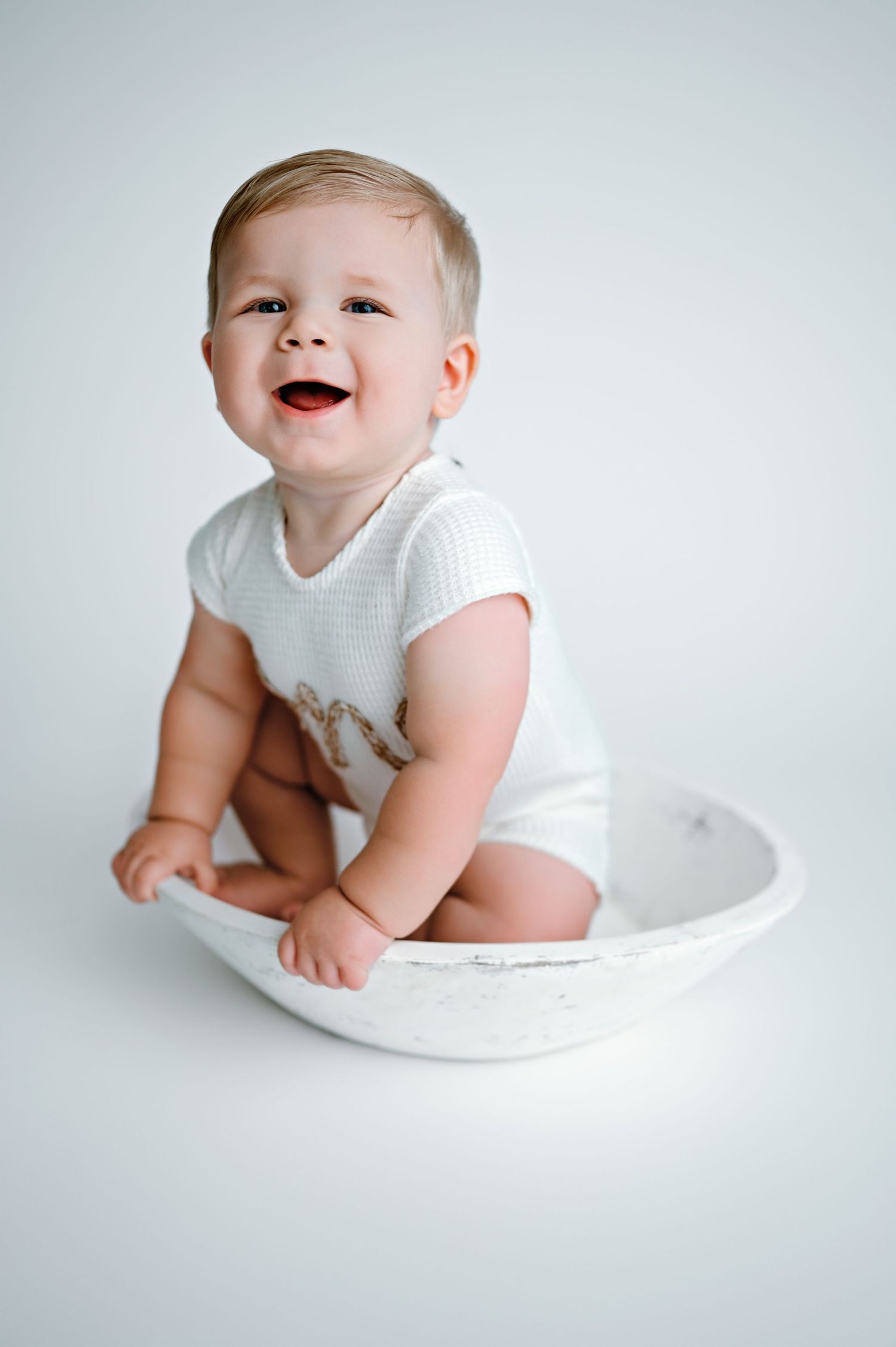 A baby wearing white clothing sits and plays in a white pillow background showing various expressions of joy.