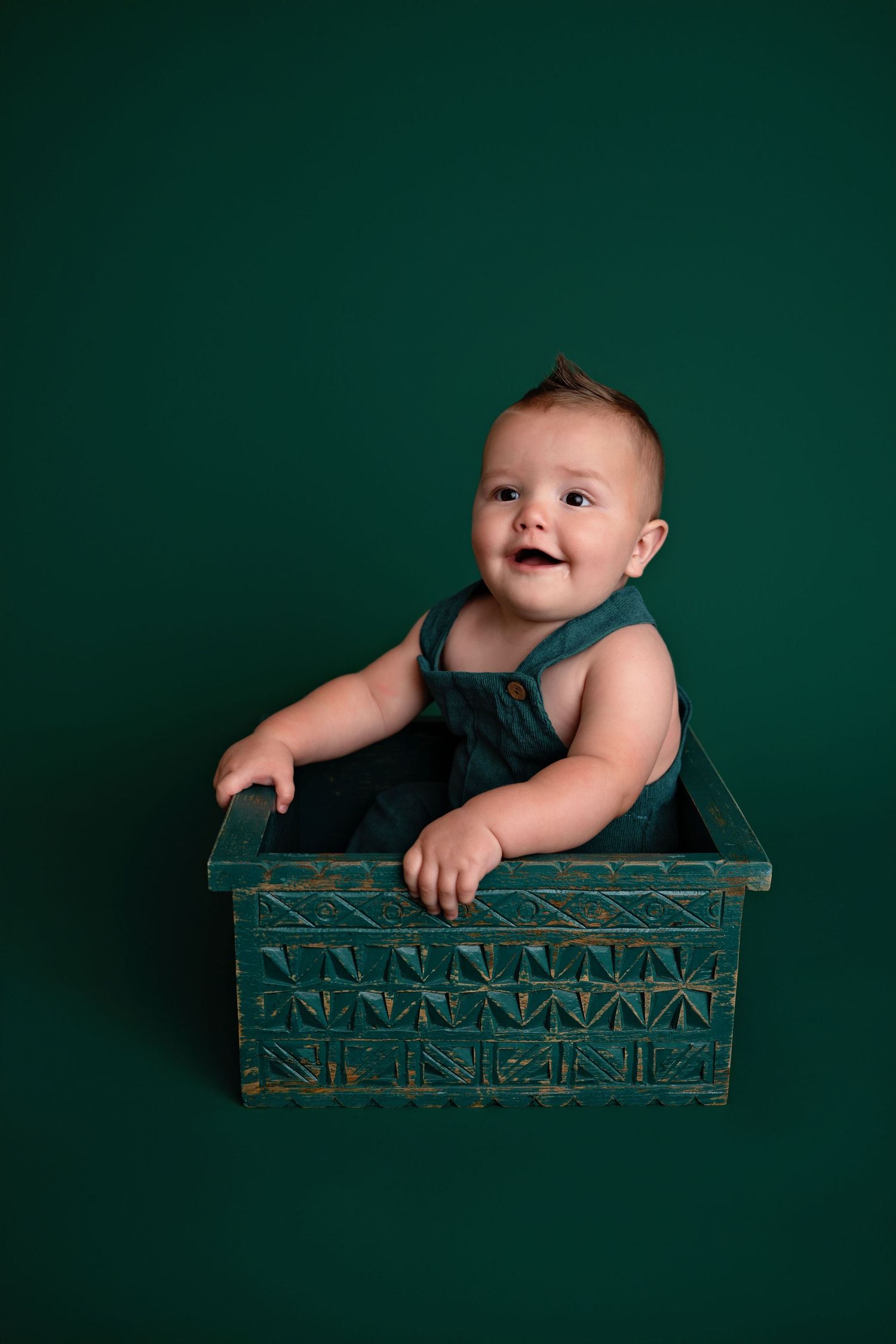 Baby sits inside vintage tin box against deep green backdrop in series of professional studio portraits.